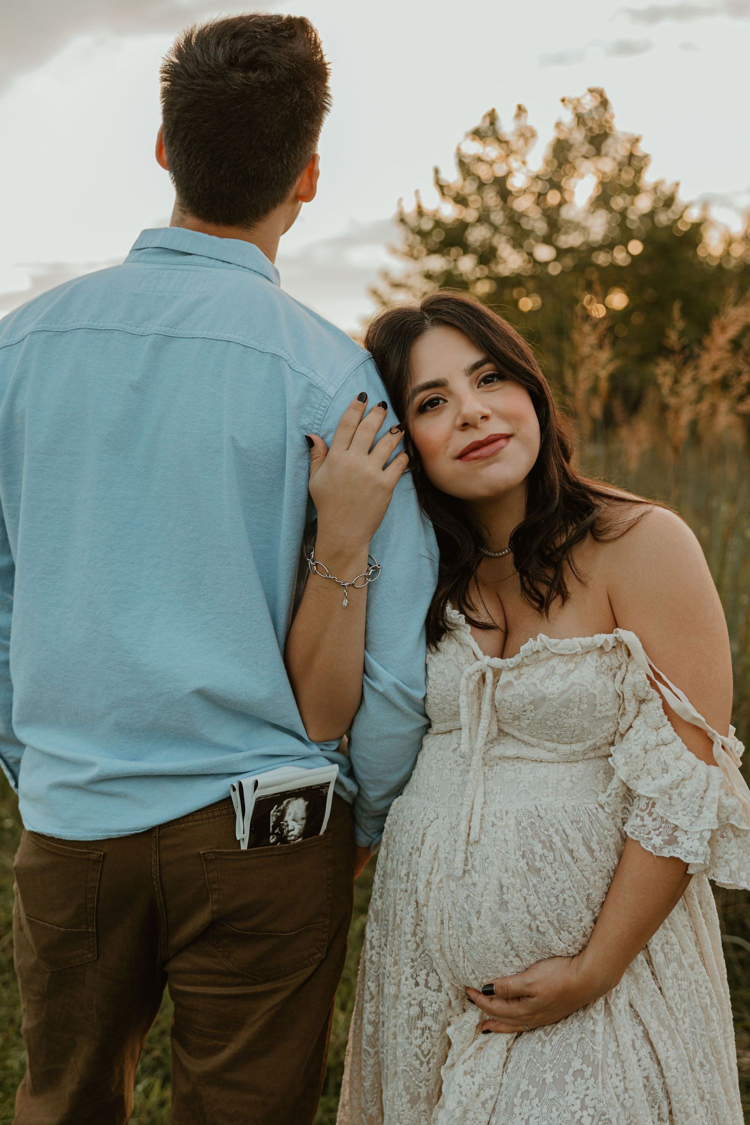 Pregnant woman in white lace off-shoulder dress smiles at camera while standing with partner outdoors at sunset.