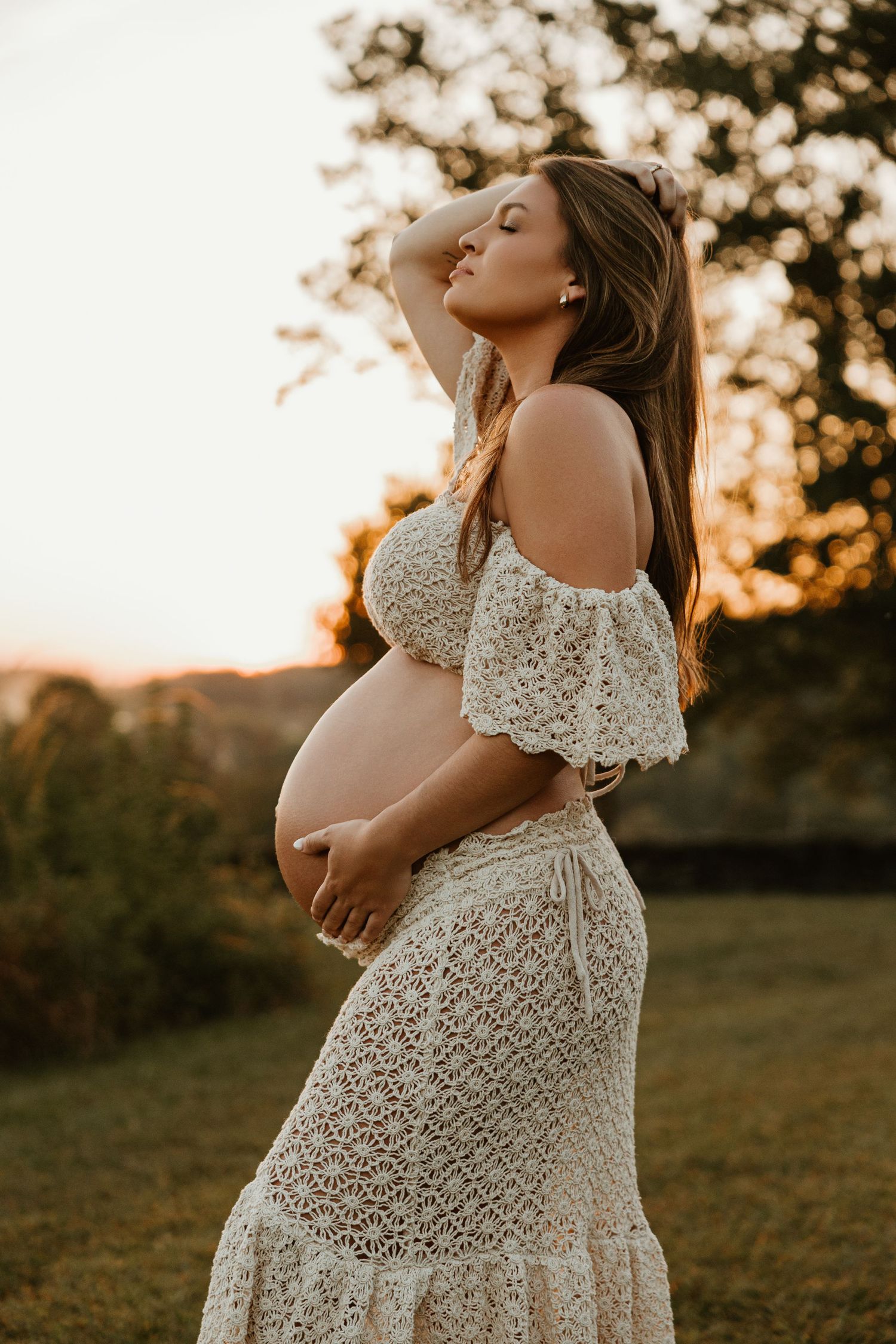 Side view maternity portrait in white lace dress during sunset with bokeh background.