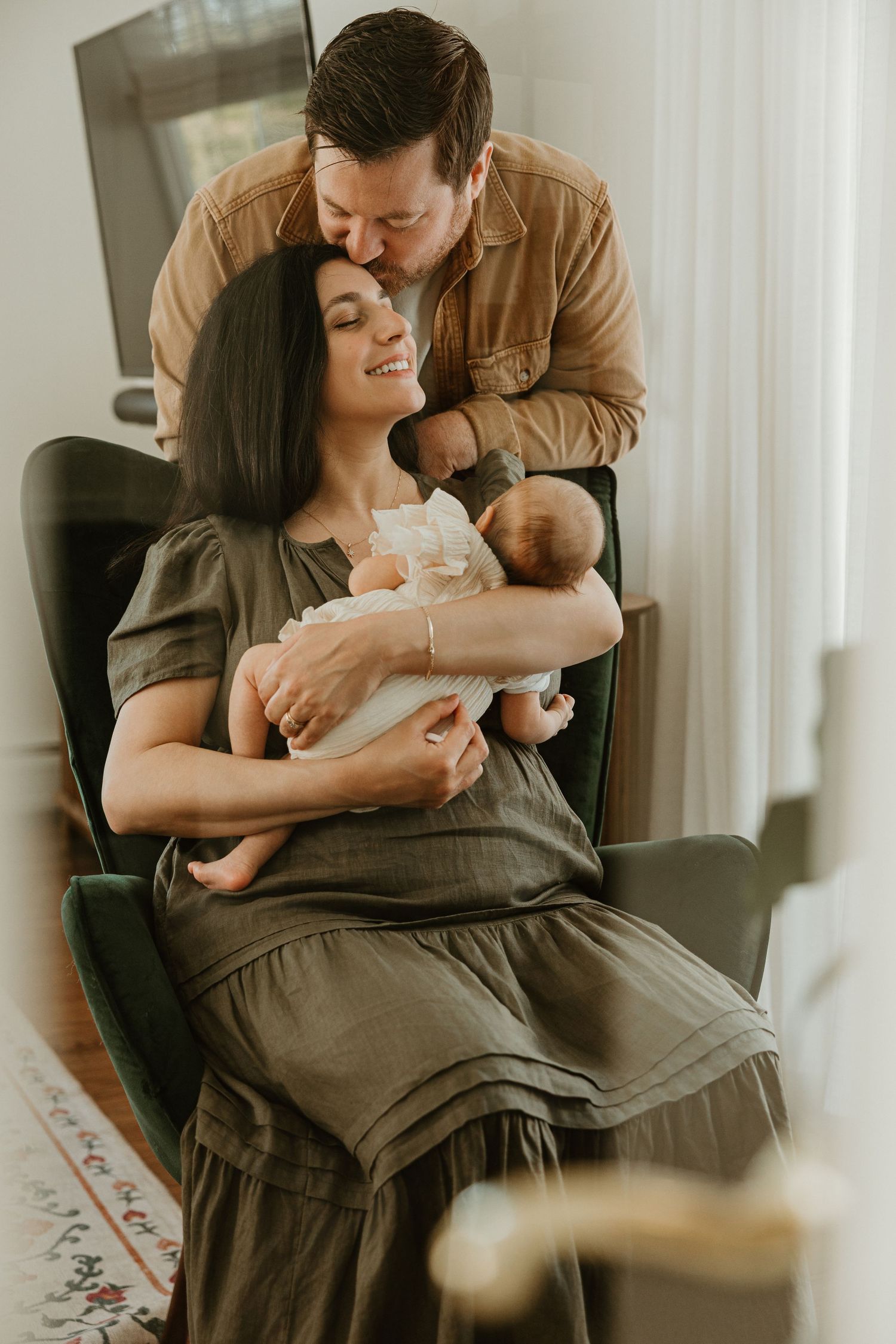A happy couple shares a tender moment with their newborn baby, as the father kisses the mothers head while she sits in a chair.