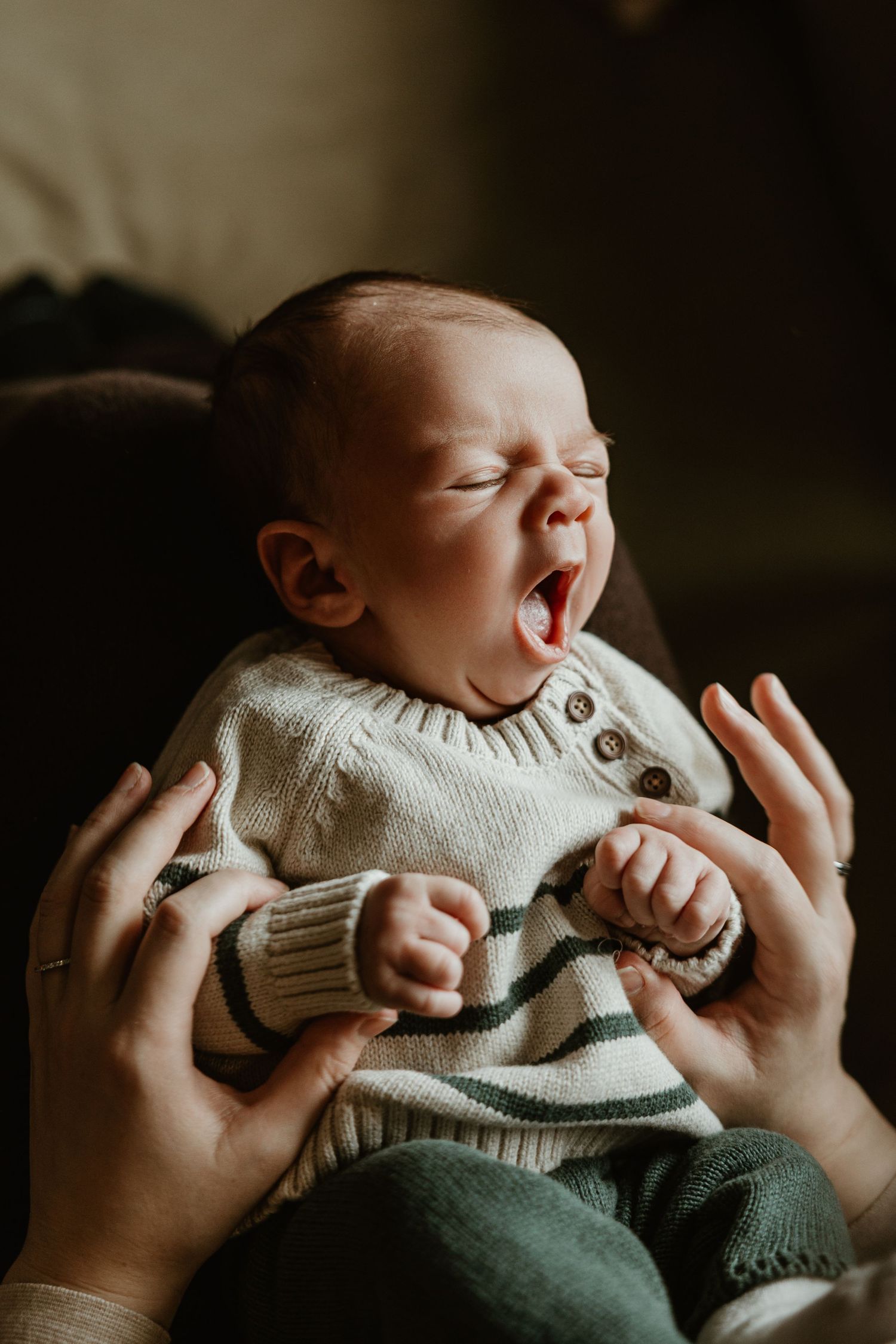 Newborn baby wearing cream sweater and green pants being held by gentle hands in moody lighting.