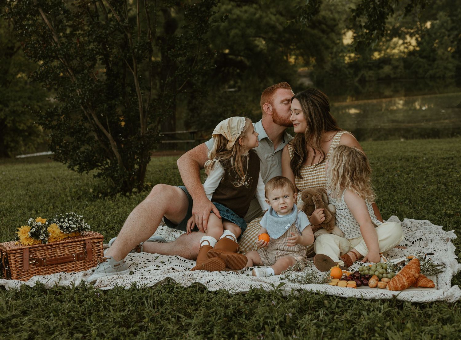 Family enjoying a picnic on a blanket in a lush green park with a wicker basket and fresh fruit nearby.
