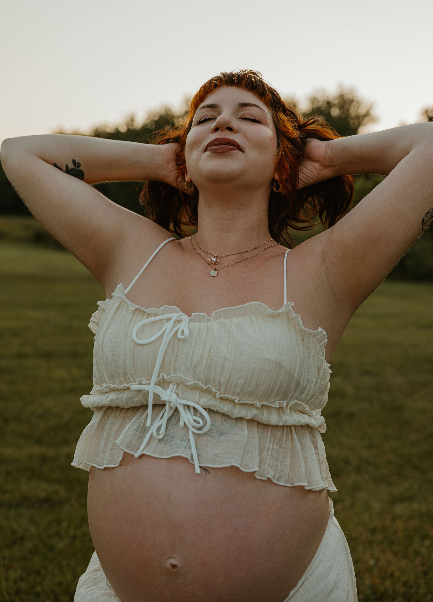 Pregnant woman in white crop top poses outdoors at sunset with hands behind her head, smiling serenely.
