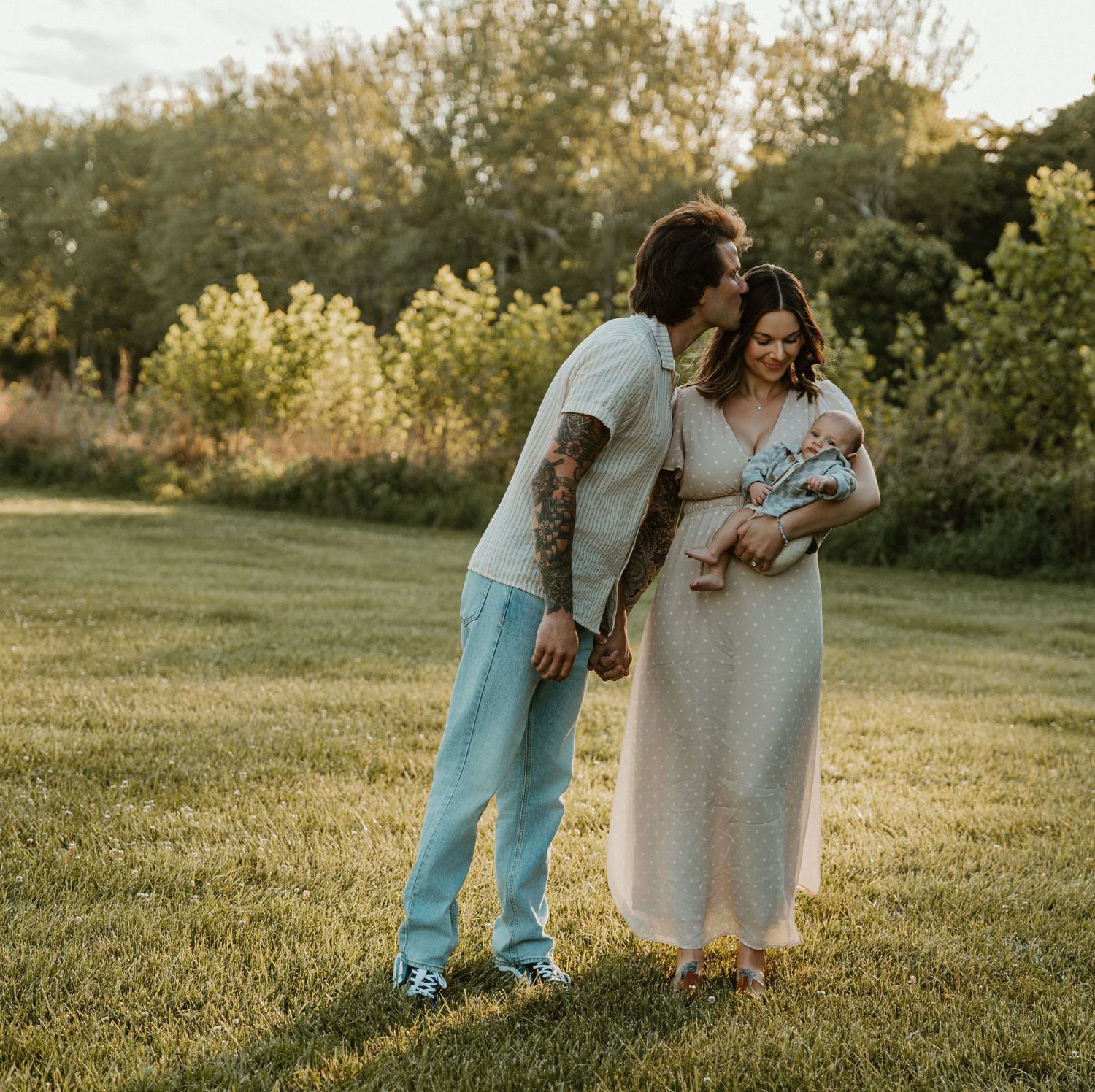 A couple in a sunlit park, man with tattoos in blue pants kisses woman in pink maxi dress holding their newborn baby.
