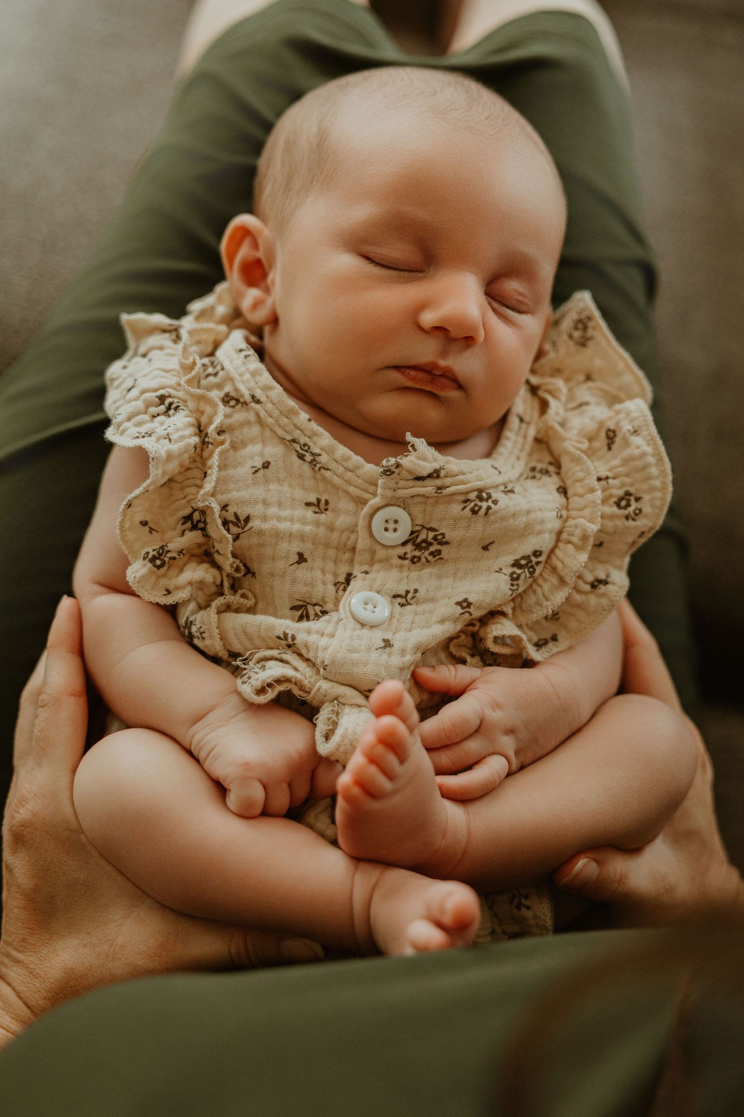 Sleeping newborn baby in floral ruffle onesie cradled in parent's arms.