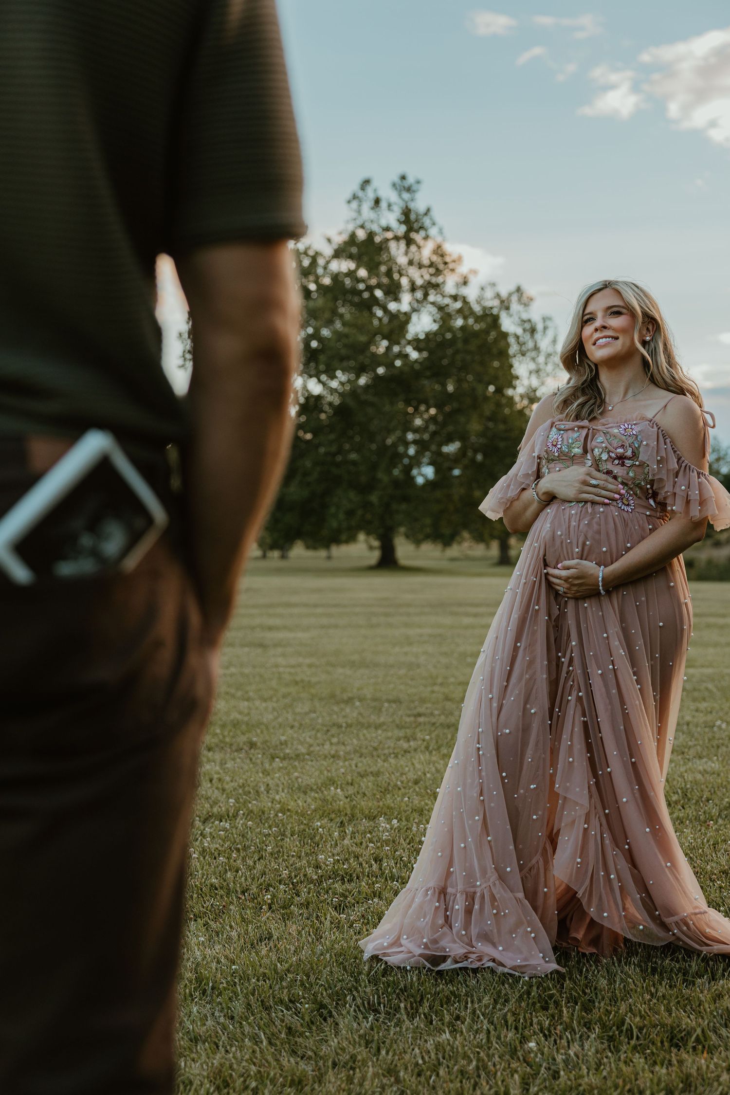 Pregnant woman in elegant blush tulle gown smiles outdoors at golden hour, holding baby bump in a lush green park.