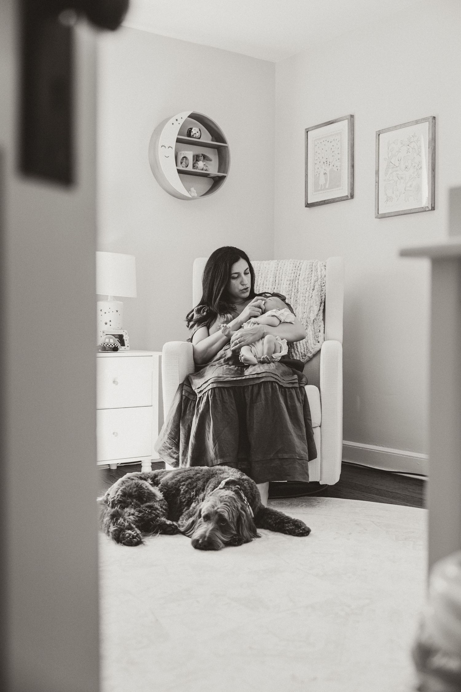 Black and white photo of a mother sitting in a nursery chair holding her newborn baby with a dog resting at her feet.