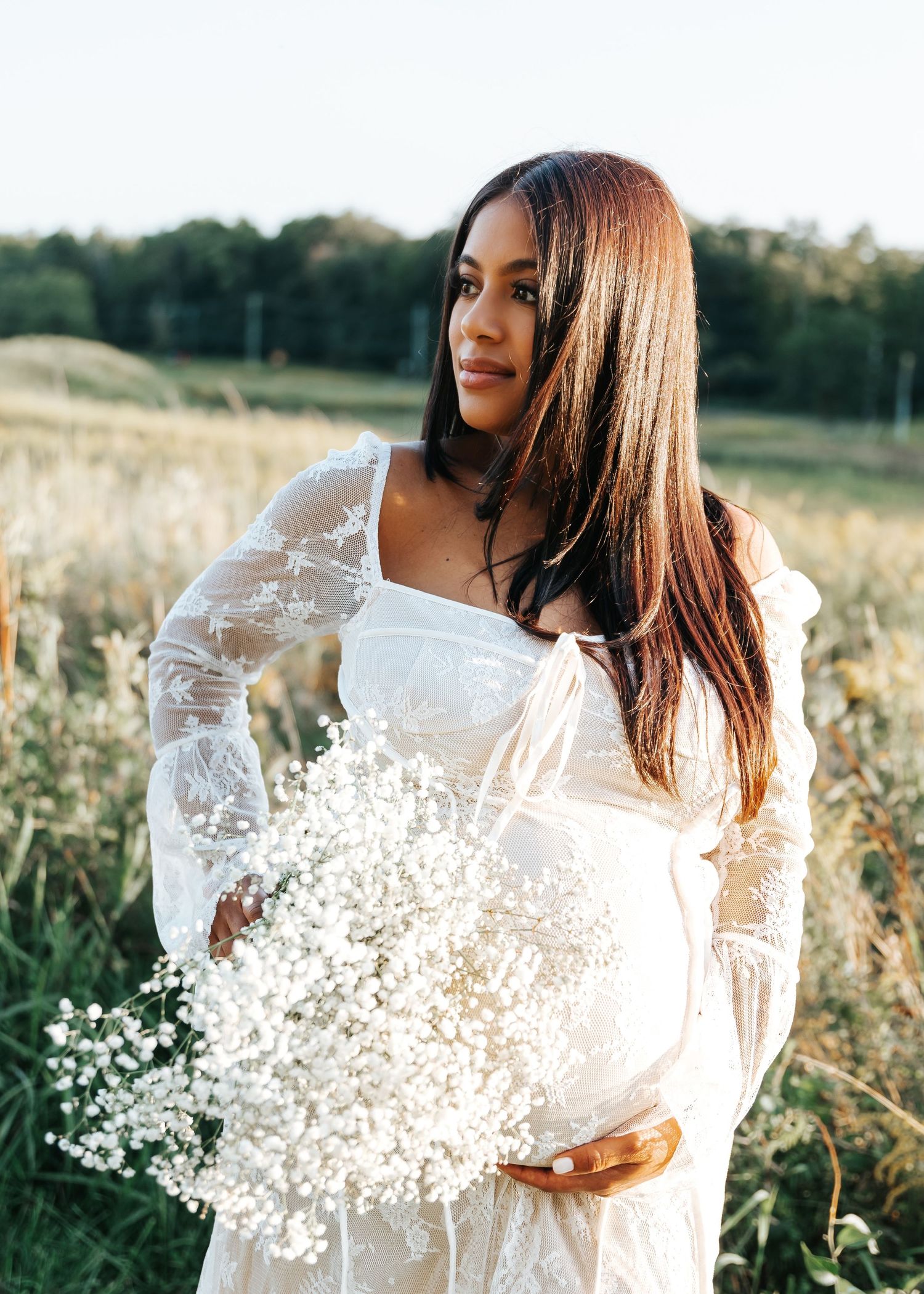 A woman in white lace holds baby's breath flowers in a golden field at sunset.