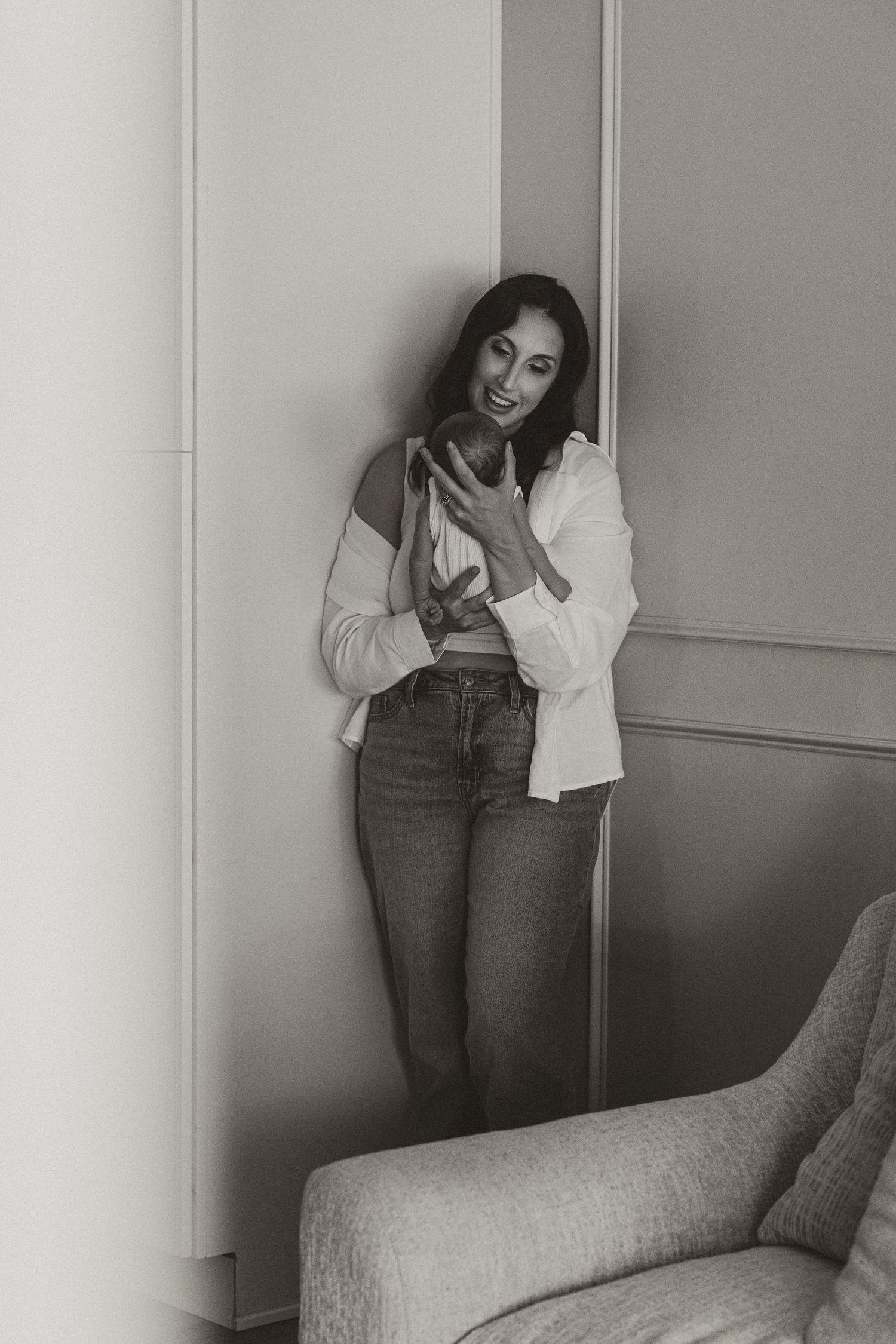 Black and white photo of a smiling woman in jeans and white shirt leaning against a wall holding a phone.
