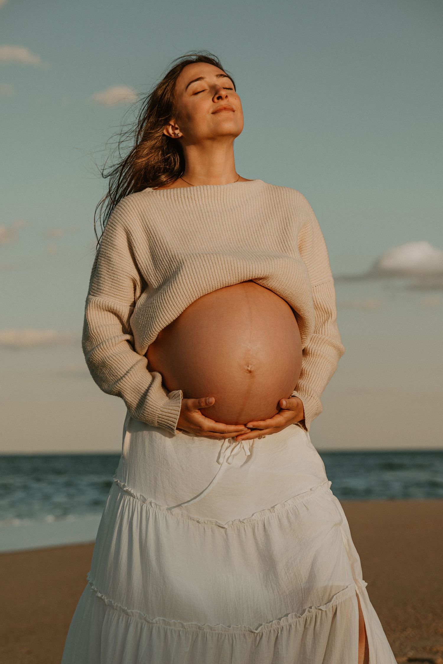Pregnant woman in cream sweater and white skirt cradling baby bump on beach with eyes closed at golden hour.