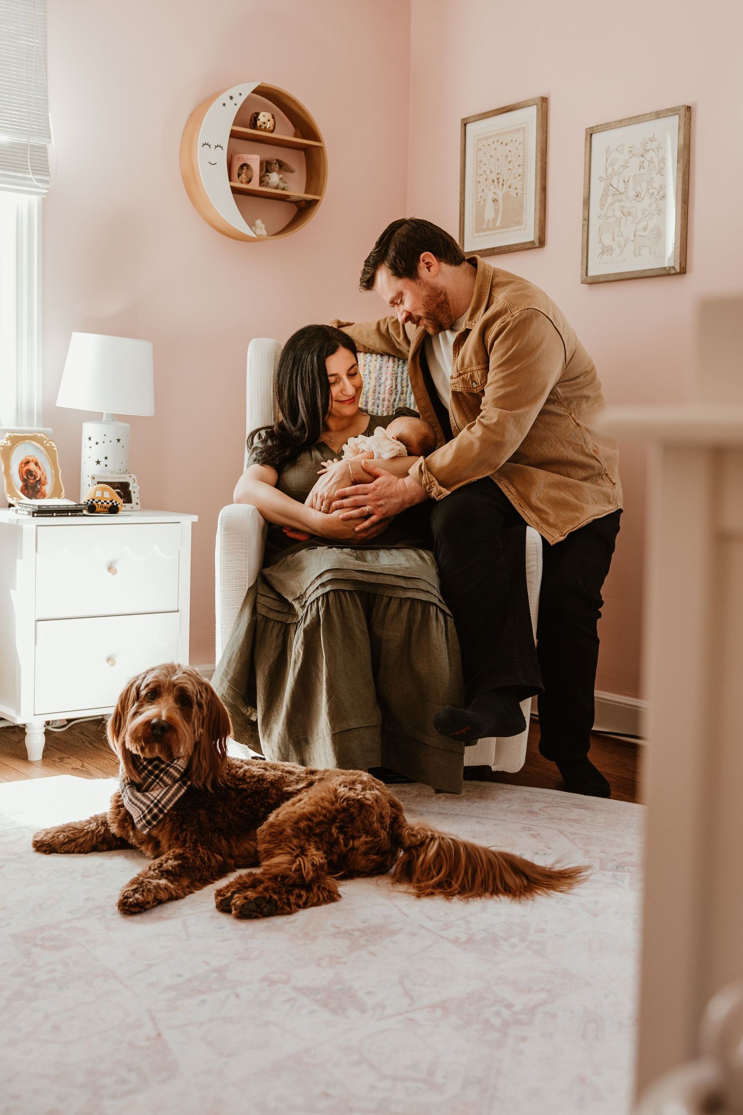 A family with a newborn sits in a pink nursery, with a fluffy brown Goldendoodle dog lying on the floor nearby.