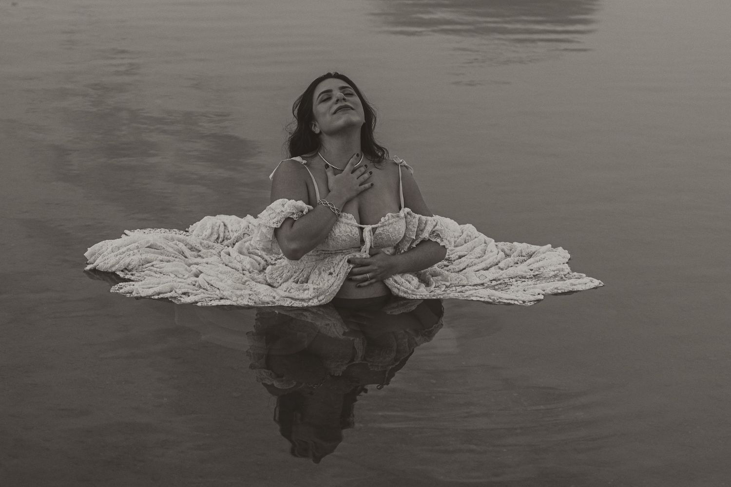 Black and white photo of a woman in a white dress sitting in calm water, gazing upward peacefully.