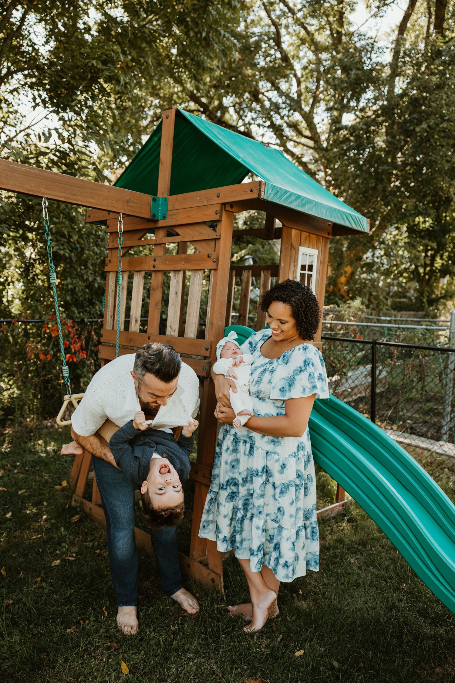 Couple playing and laughing together on a backyard swing set with wooden playhouse in background.
