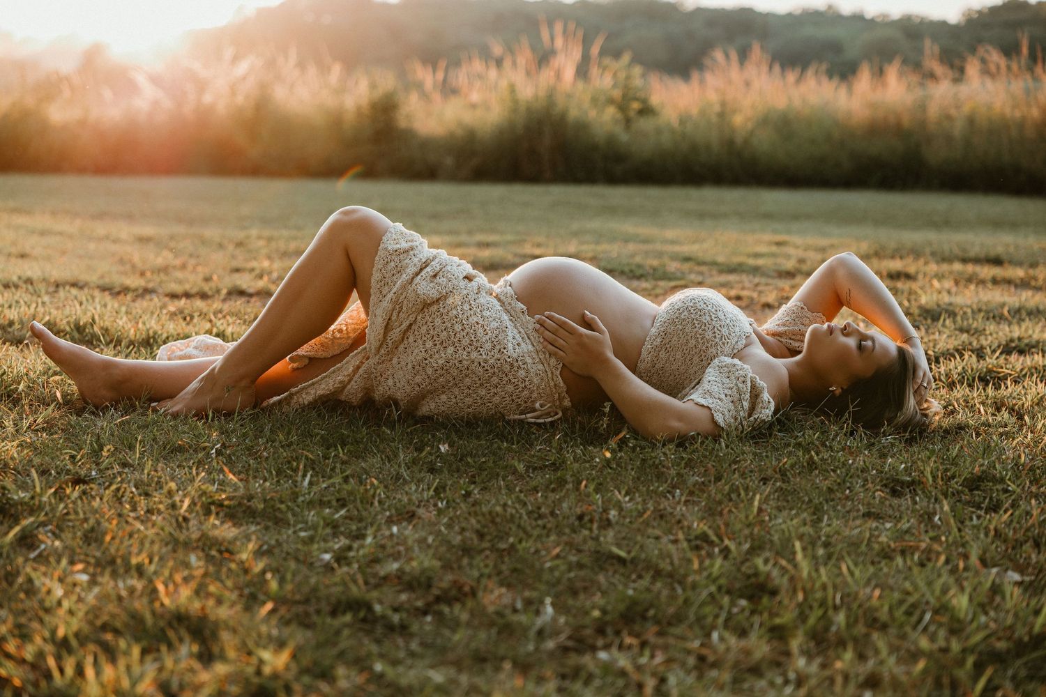 Maternity portrait of person in white dress lying on grass during golden hour sunset.