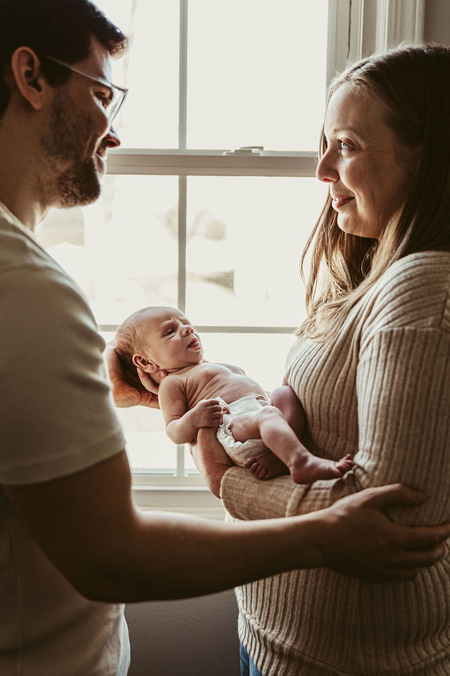 Candid sequence of parents sitting by window sharing tender moments with their newborn.