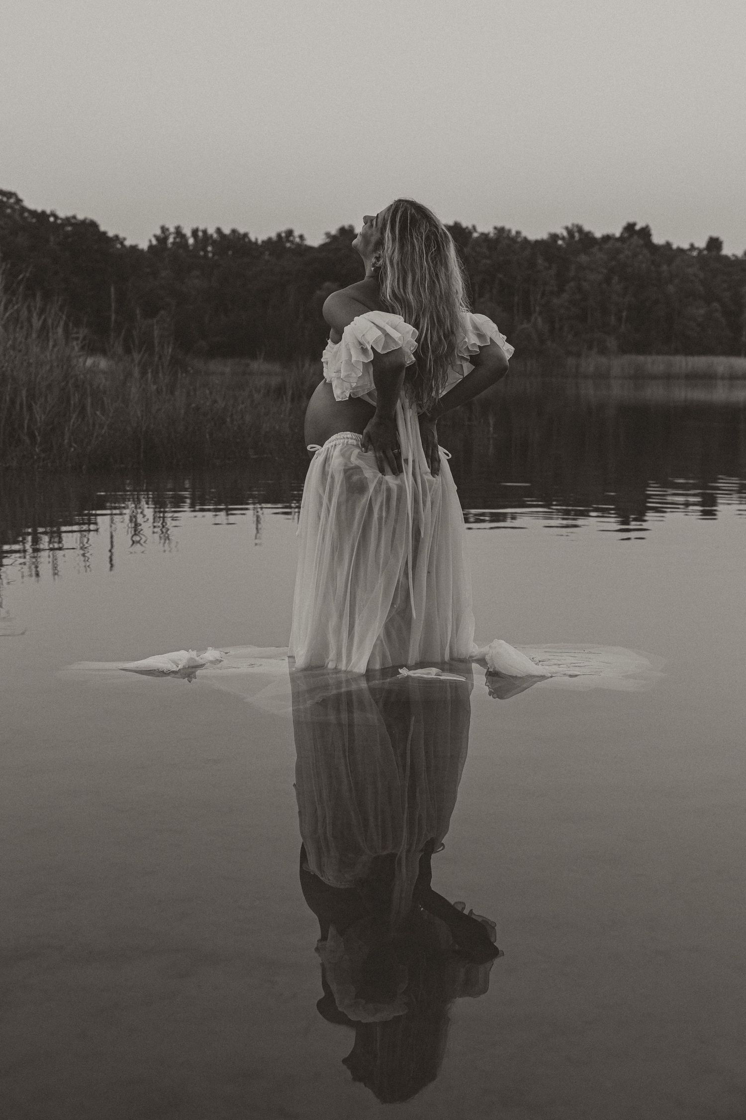 Black and white photo of a woman in a white dress standing in a calm lake with her reflection visible in the water.