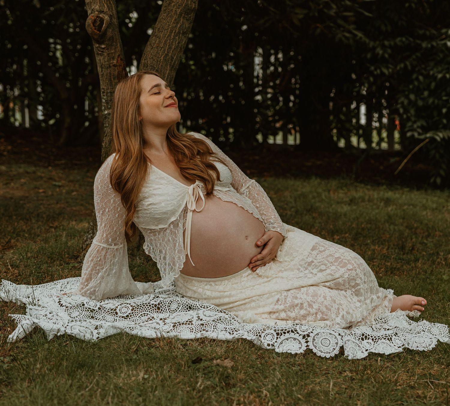 Pregnant woman in white lace dress sitting on grass, eyes closed, cradling baby bump outdoors.
