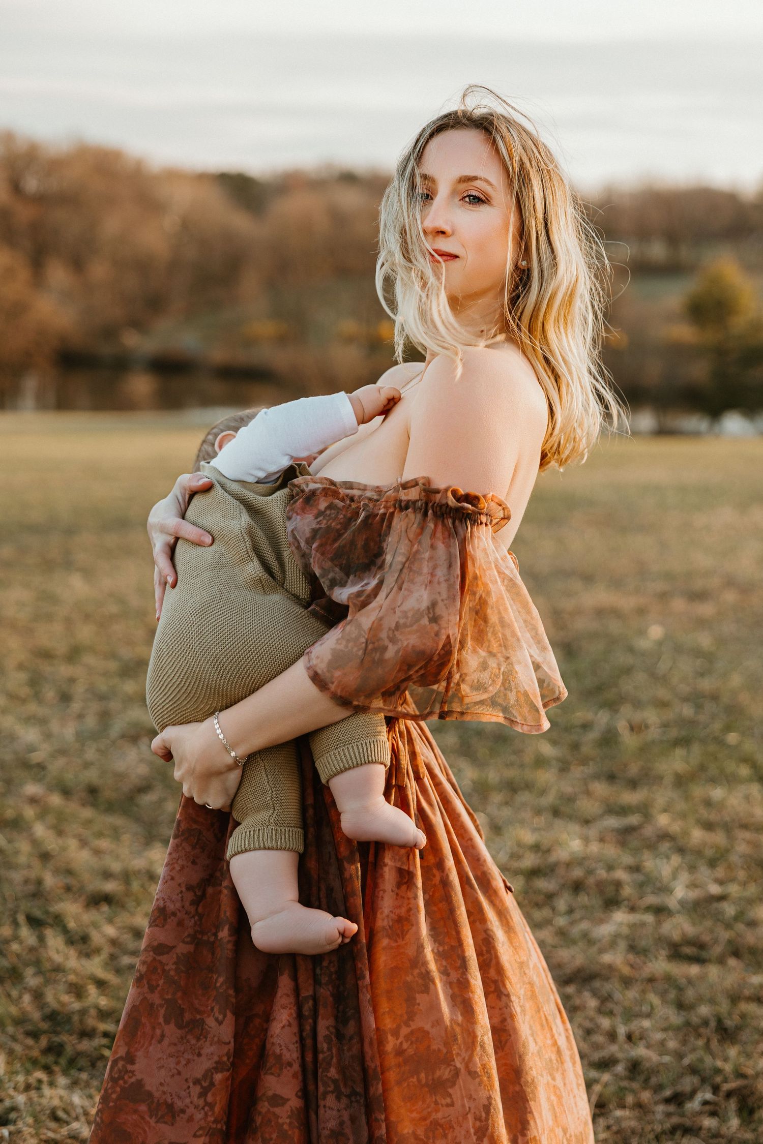 A blonde woman in a rust floral off-shoulder dress holds her baby outdoors in a golden field at sunset.