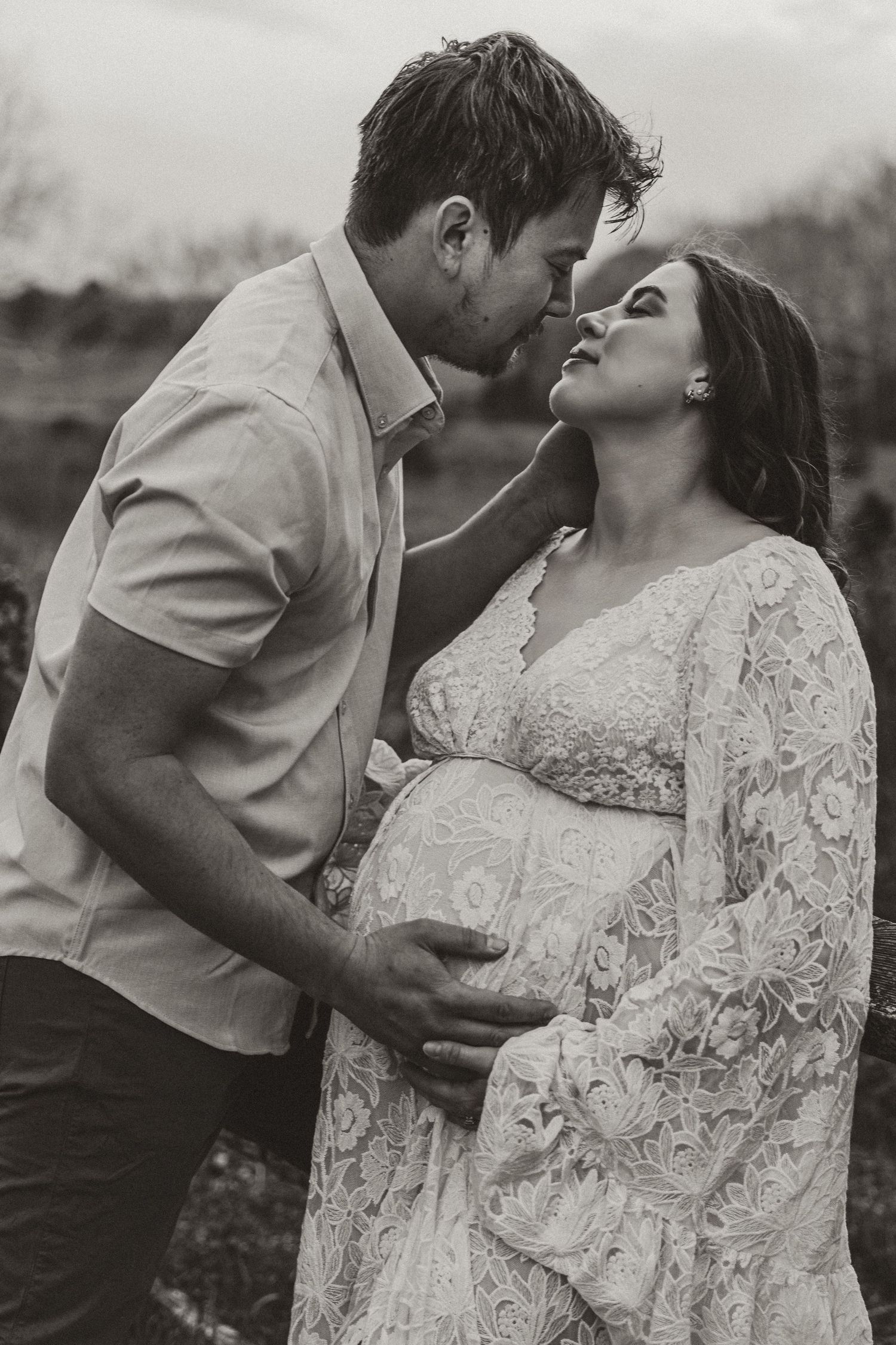 A black and white photo of a couple sharing an intimate moment, the woman in a lace maternity gown.