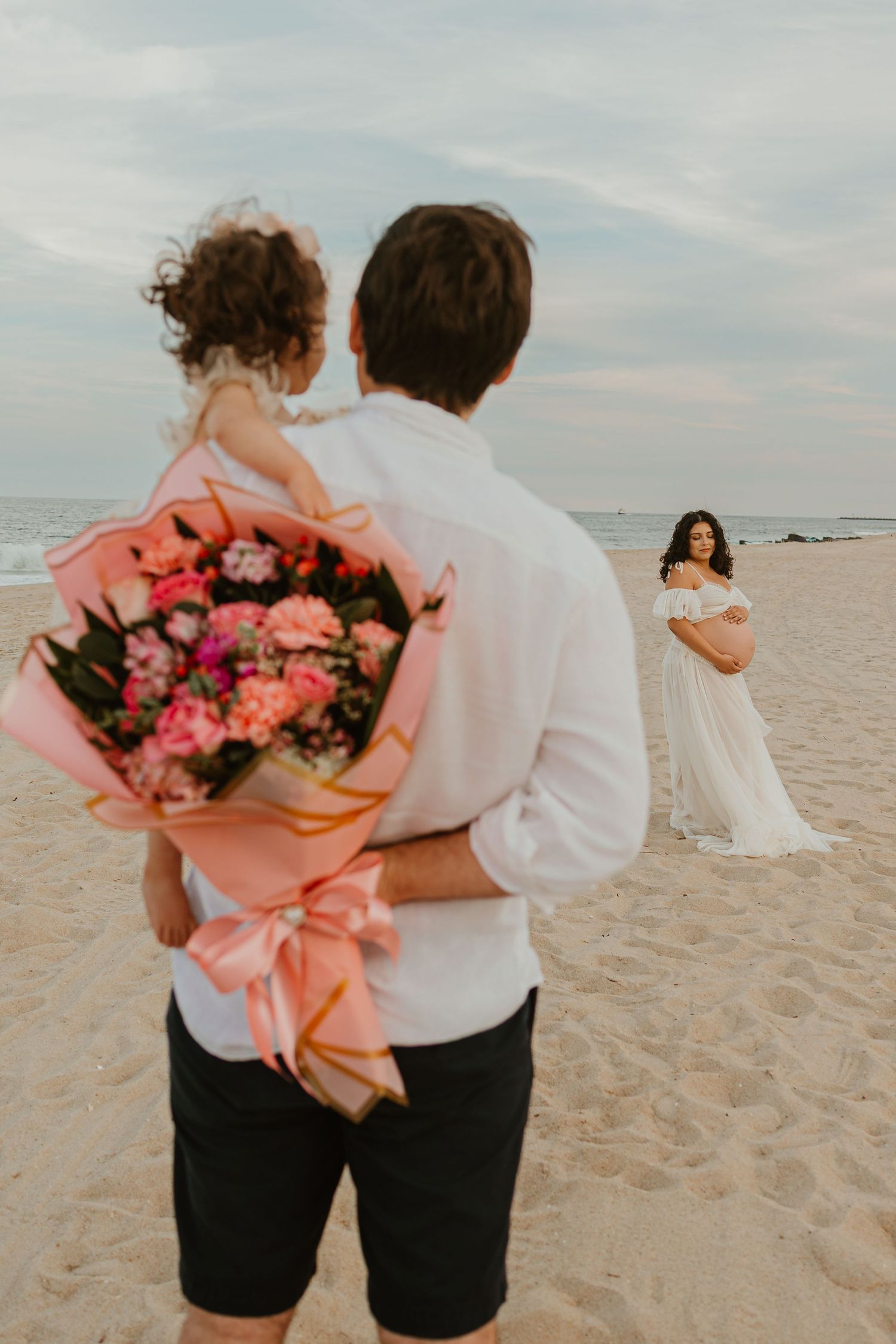 Man holding flowers and child on beach with pregnant woman in white dress during maternity photoshoot.