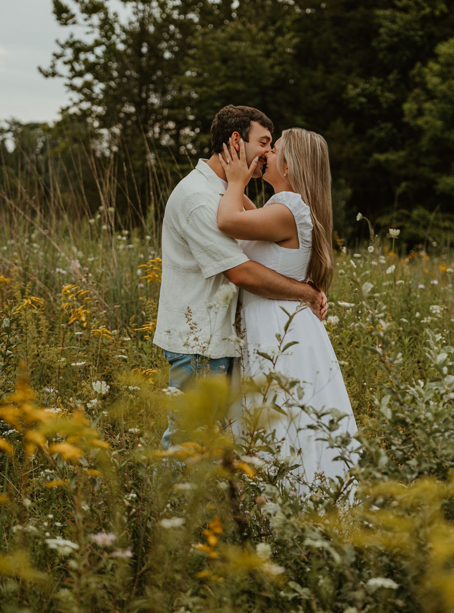 Couple sharing a romantic kiss in a wildflower meadow, dressed in white, surrounded by lush greenery.