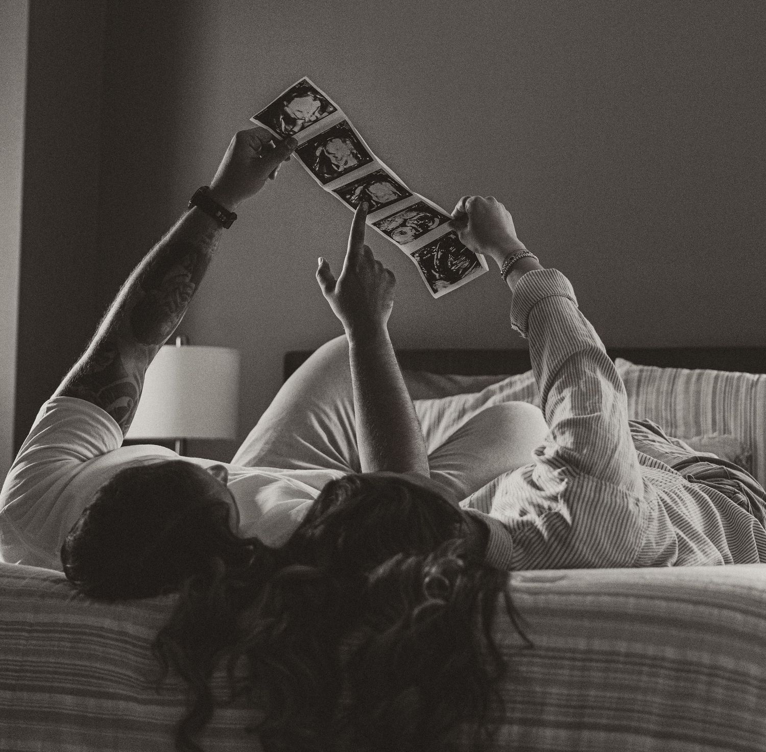 Couple lying on bed in black and white photo, holding up ultrasound images together in a tender moment.