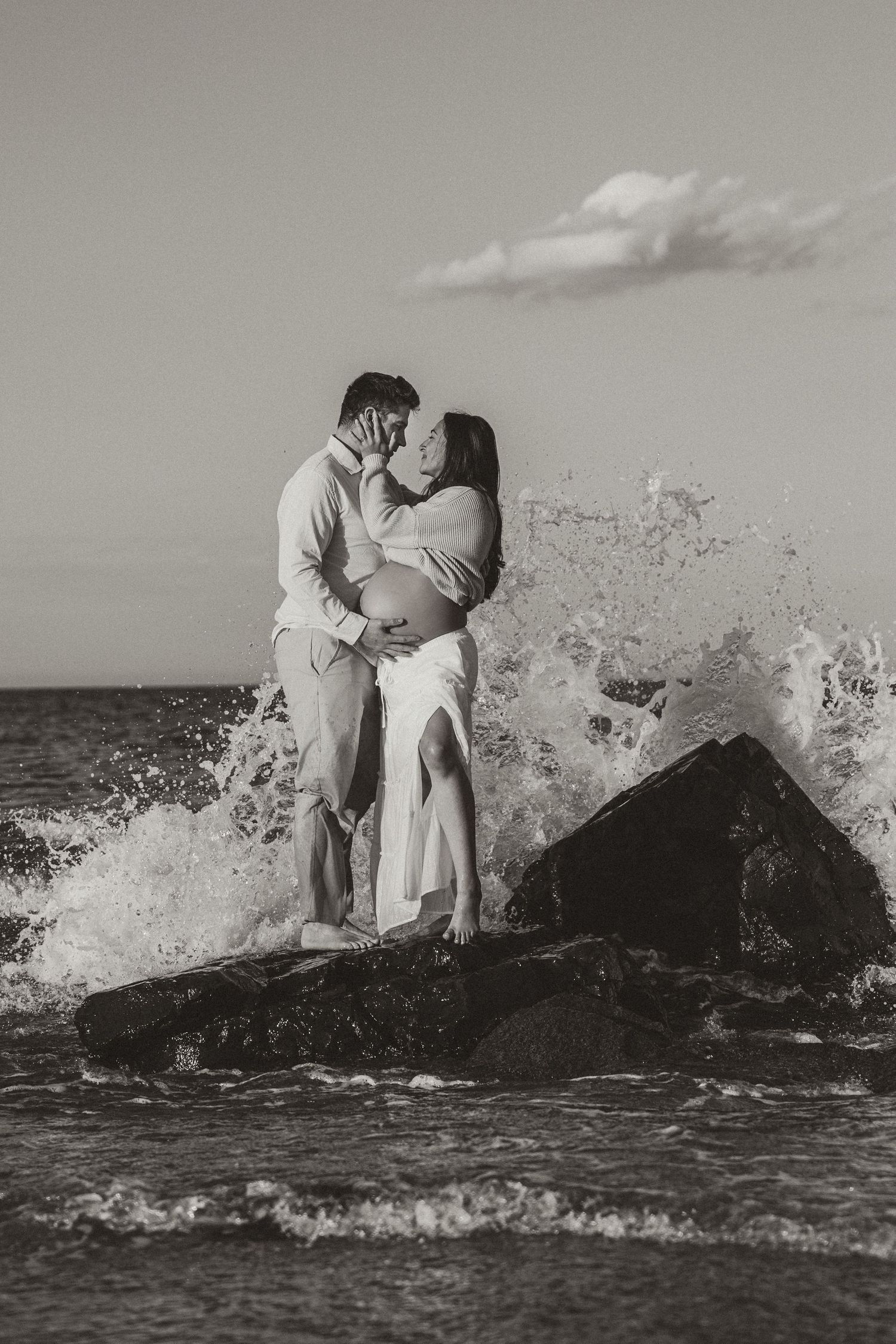 Black and white photo of a couple kissing on rocks with ocean waves splashing around them during a maternity shoot.