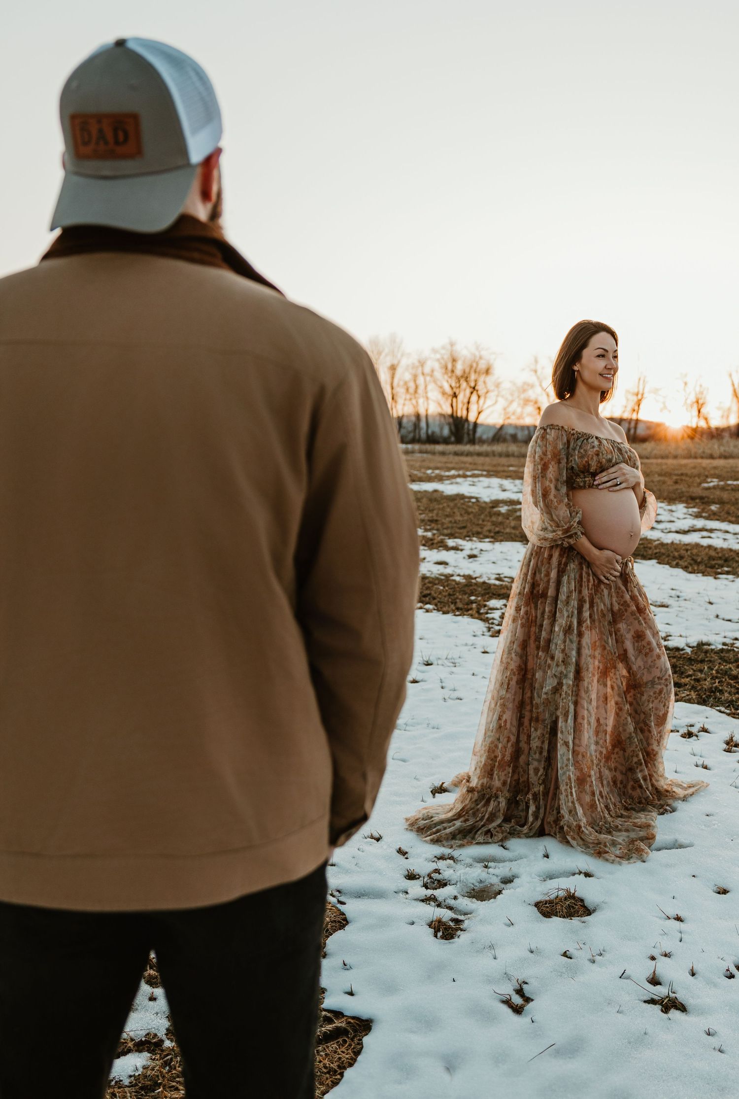 Maternity photoshoot in a snowy field at sunset with a flowing floral dress and casual winter attire.