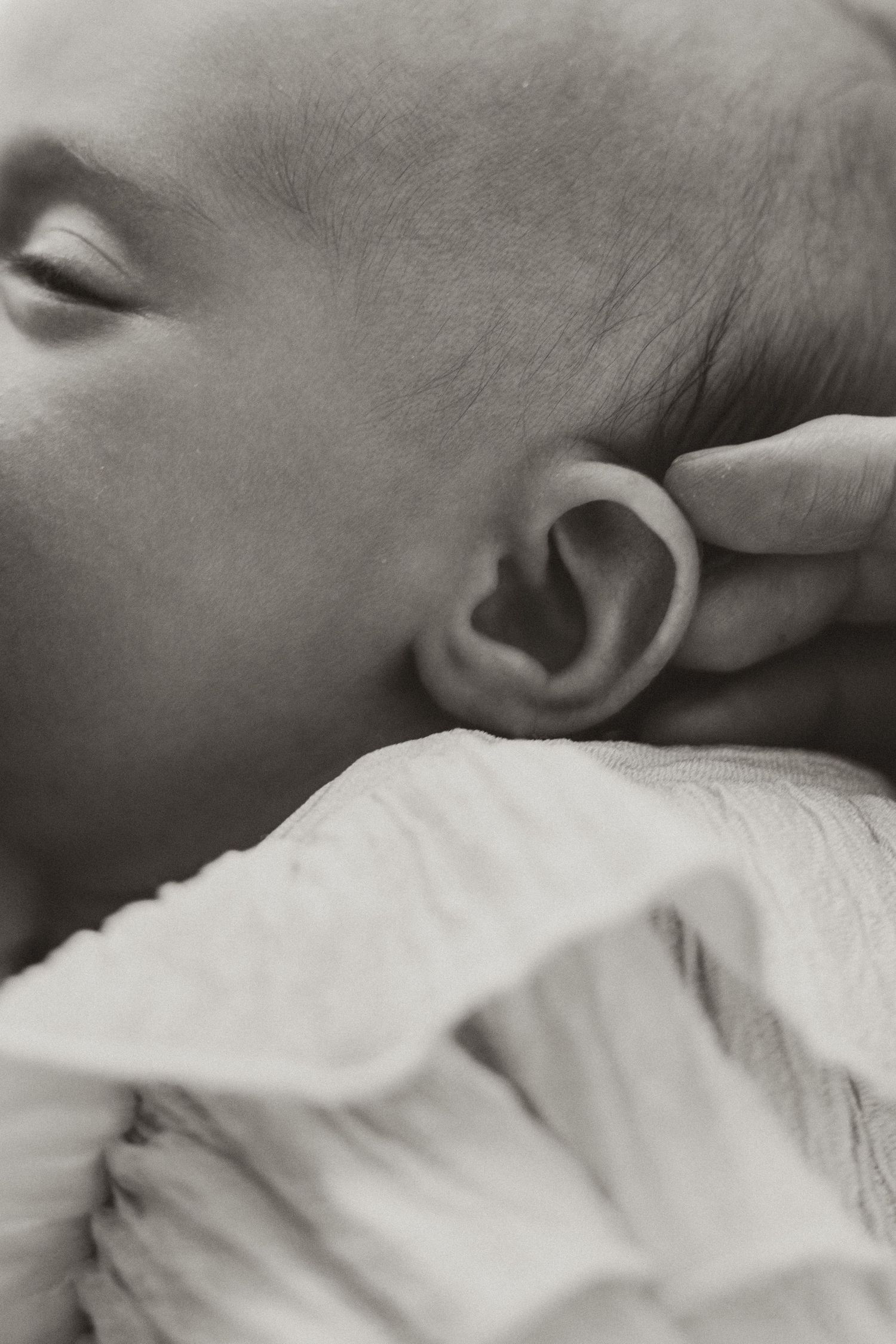 Close-up black and white photo of a sleeping newborn baby's ear and cheek resting on soft white fabric.