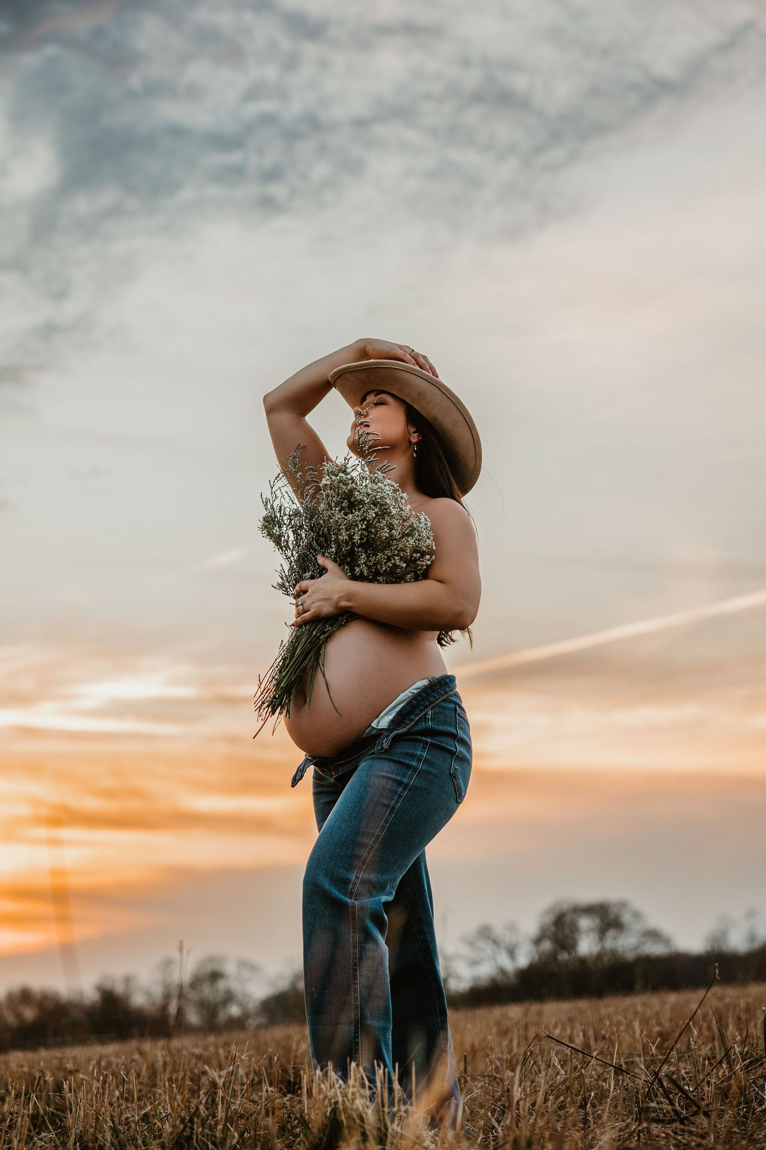 Pregnant woman in cowboy hat holding wildflowers at sunset in a field, wearing jeans with exposed baby bump.
