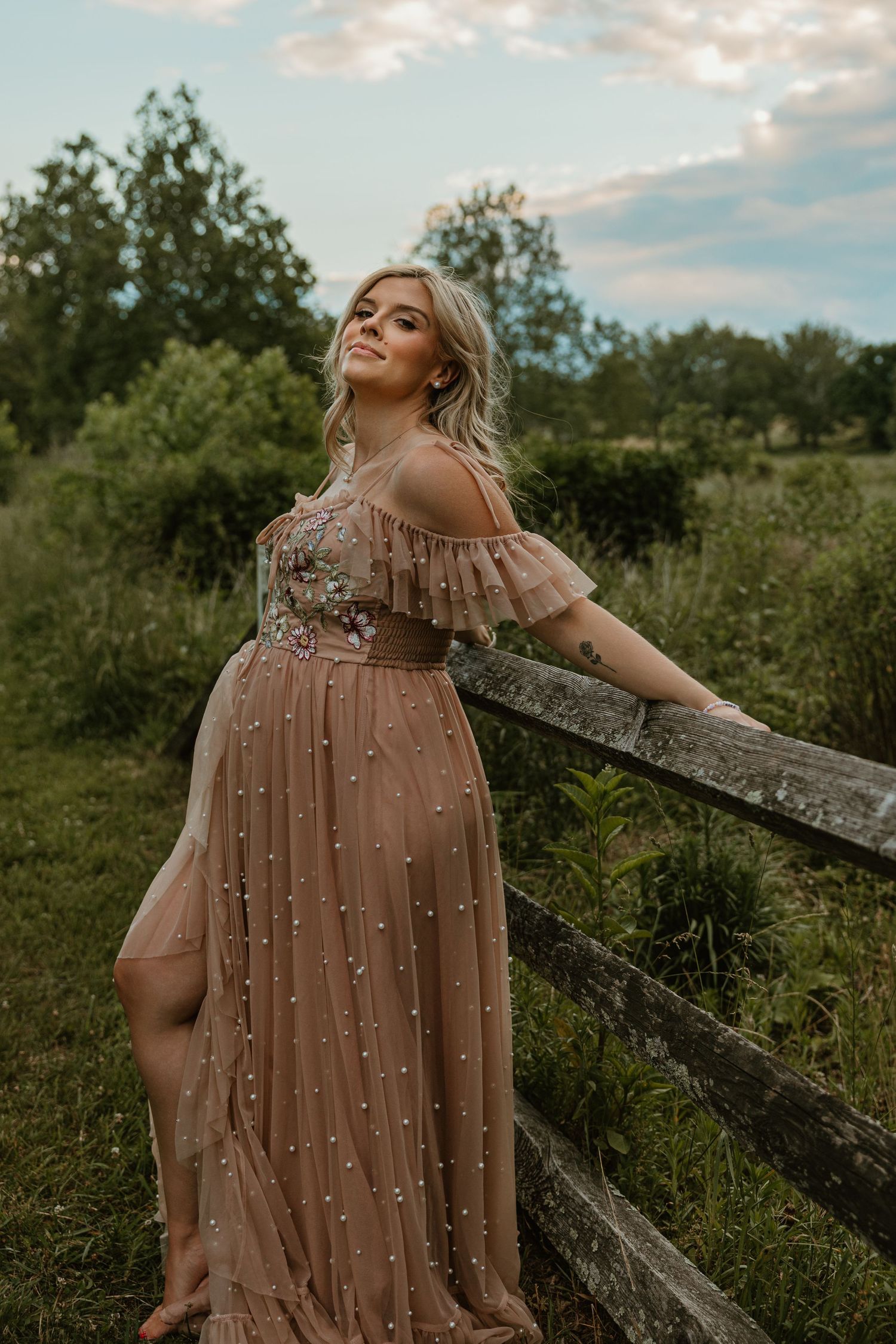 Pregnant woman in beige pearl-embellished tulle gown leaning on rustic fence in lush green field at dusk.