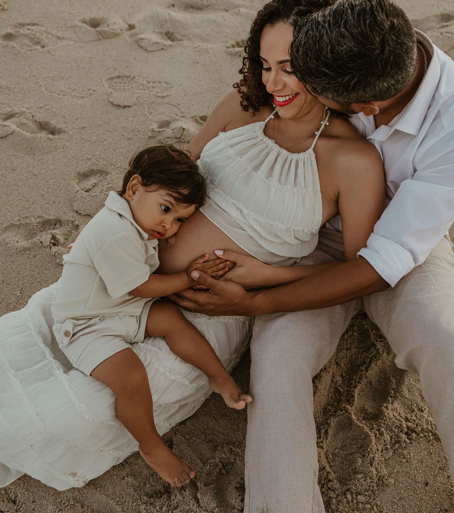 A pregnant woman sits on sandy beach with her husband and toddler, all dressed in white, touching her baby bump.