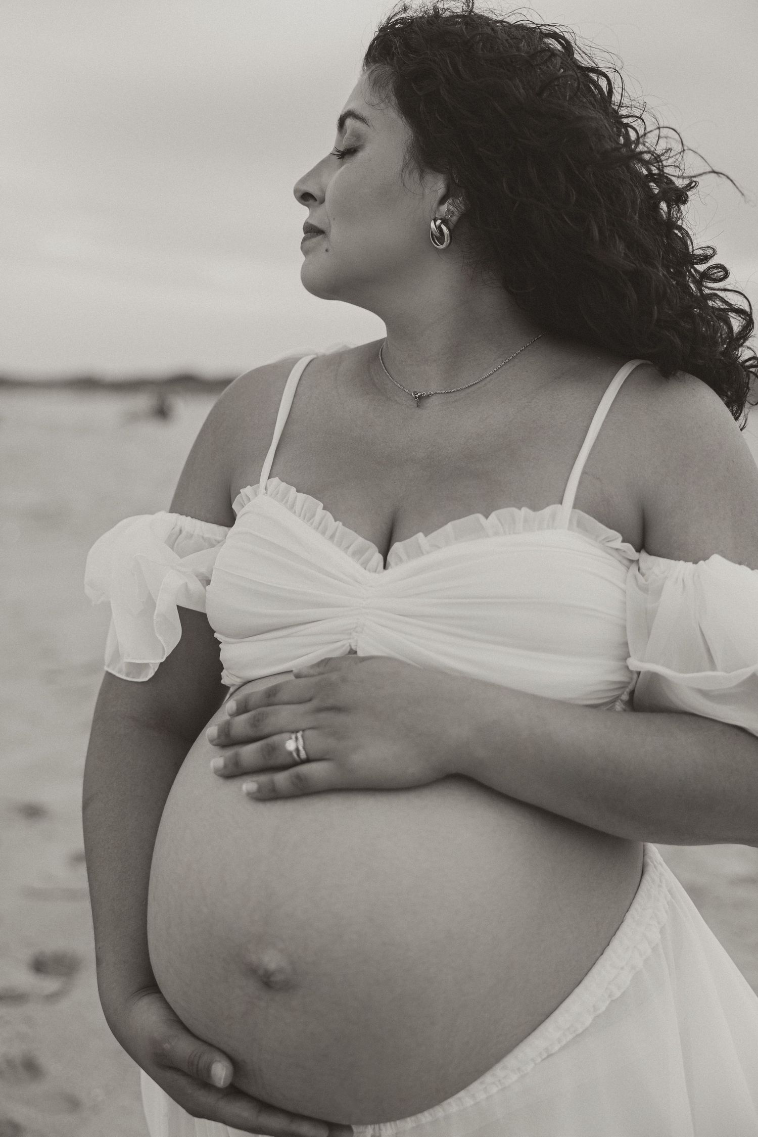 Black and white maternity photo of a pregnant woman in a white dress cradling her bare belly on a beach.