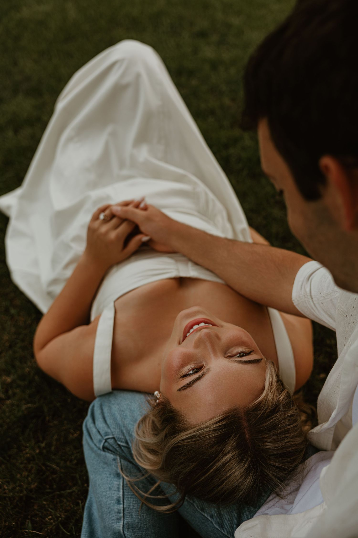 A smiling woman in white top and jeans lies on grass while a man holds her hands in a romantic outdoor moment.