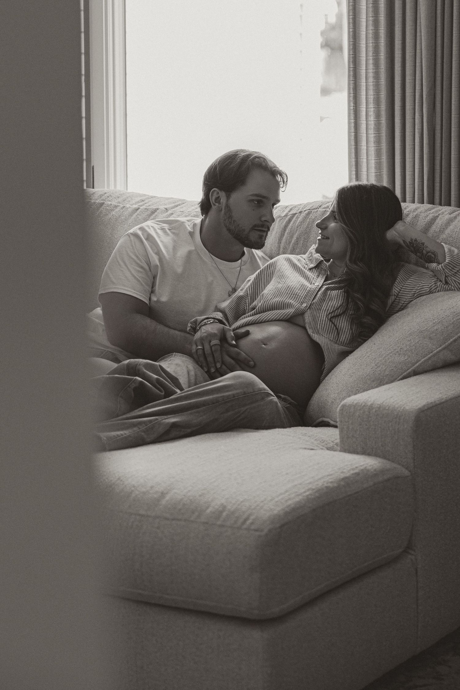 Black and white photo of a couple sitting on a couch, woman visibly pregnant, sharing a tender moment.