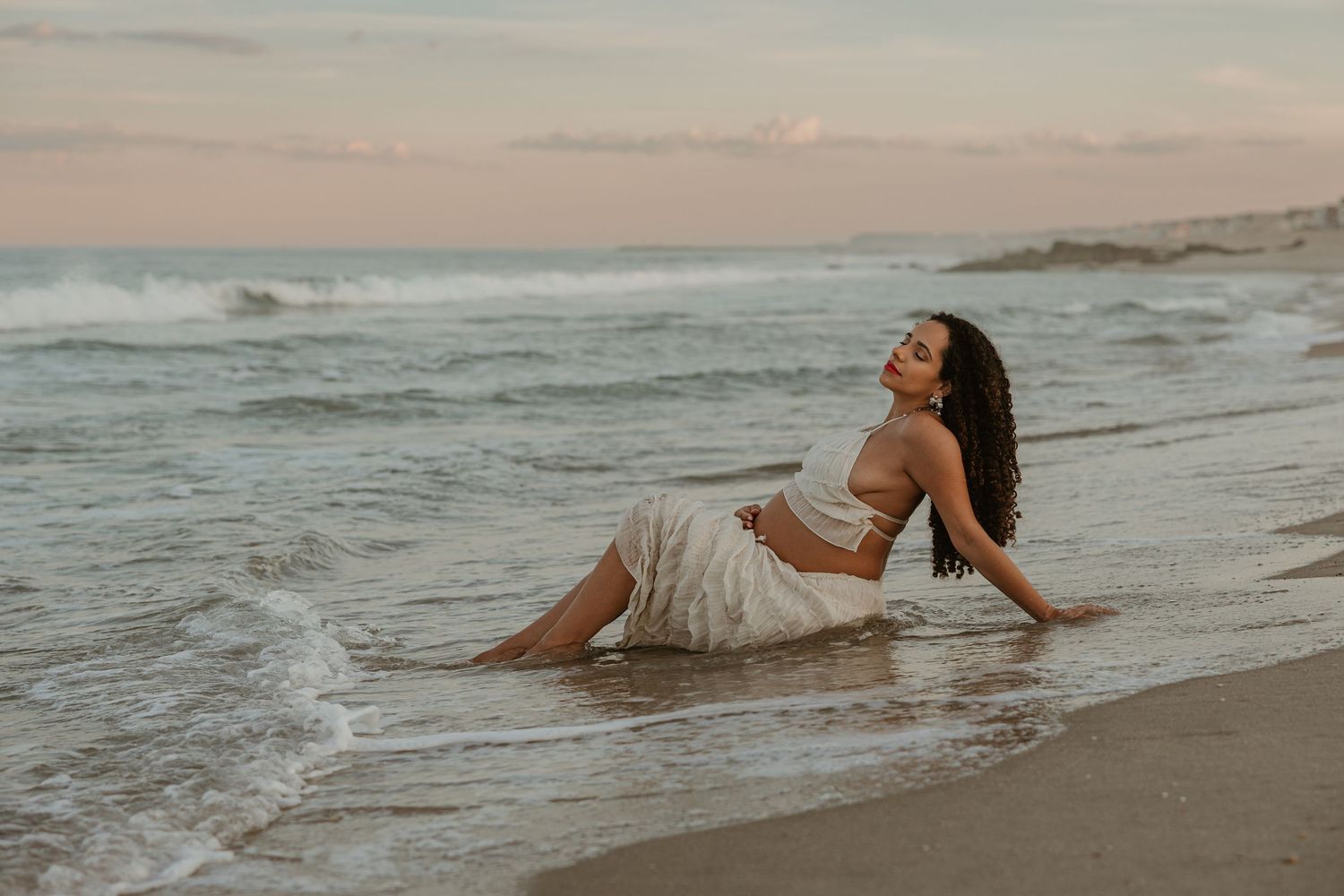 Pregnant woman in white crop top and skirt poses gracefully in ocean waves on a sandy beach at sunset.