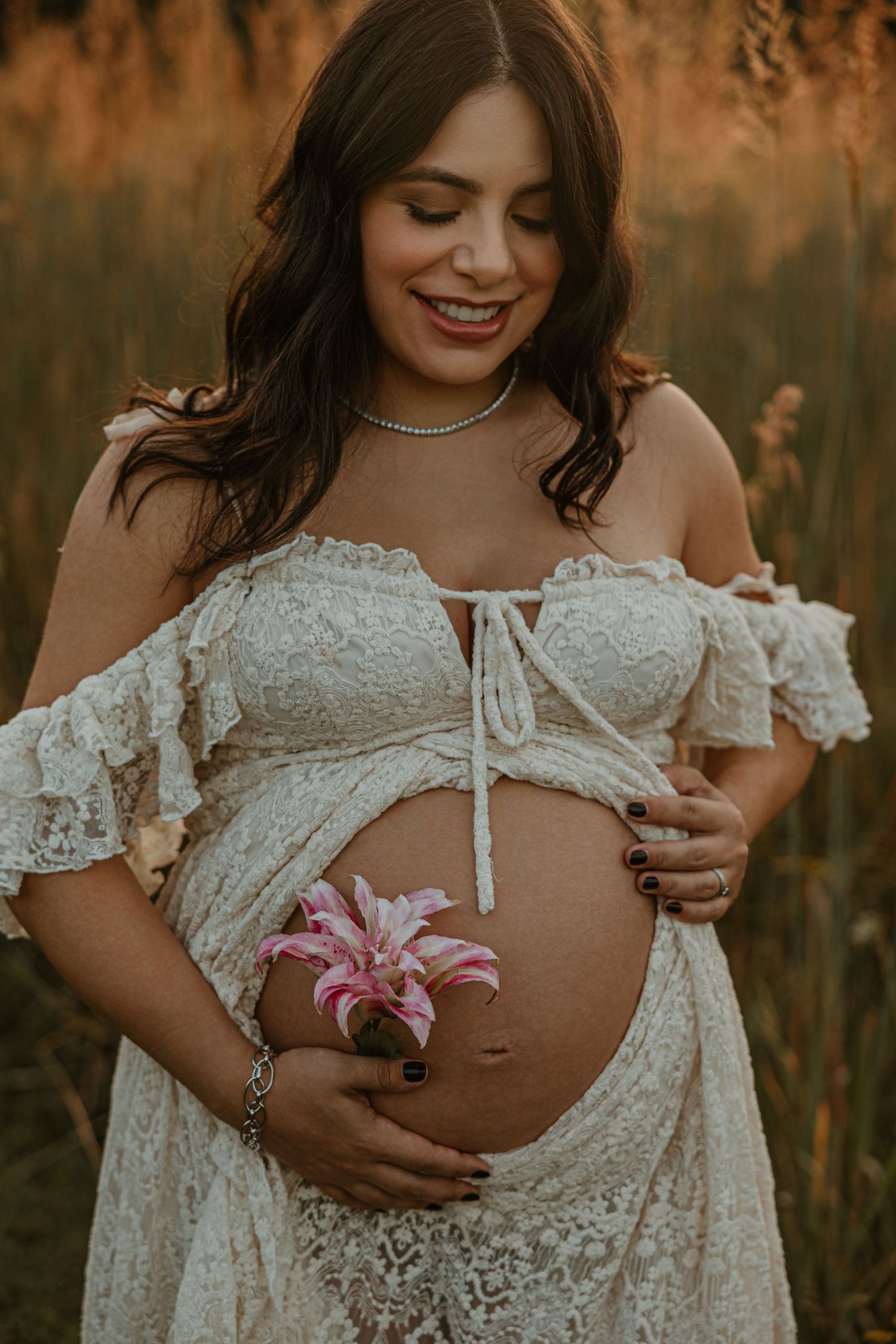 Smiling pregnant woman in white lace off-shoulder dress holding pink lily flowers in golden field at sunset.