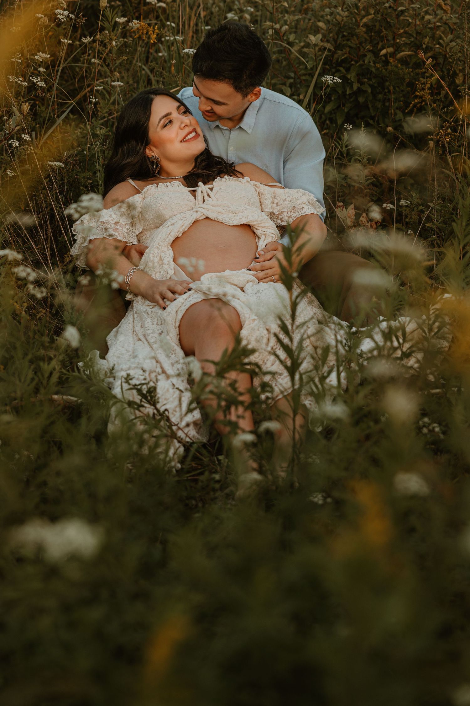 Couple sitting in wildflower field, pregnant woman in lace dress cradled by partner, golden hour maternity photo.