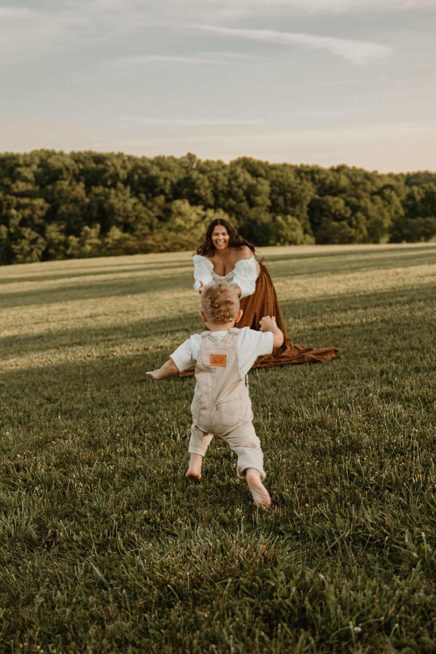 A toddler in white romper walks toward smiling mother in brown dress on lush green field at golden hour.