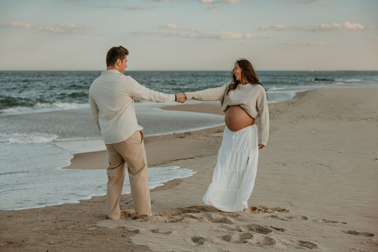 Couple holding hands on sandy beach during maternity photo shoot at sunset.