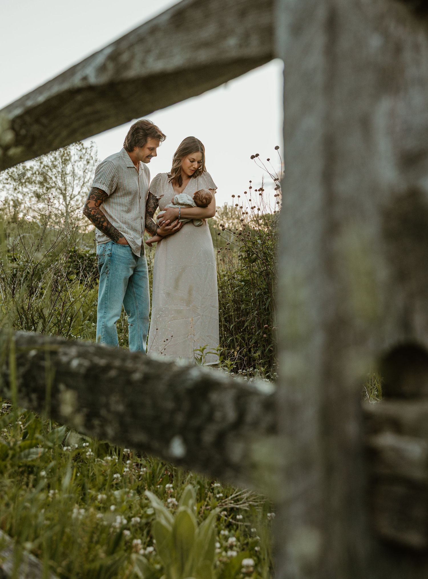 Couple stands in wildflower field framed by rustic wooden fence, tenderly holding their newborn baby outdoors.