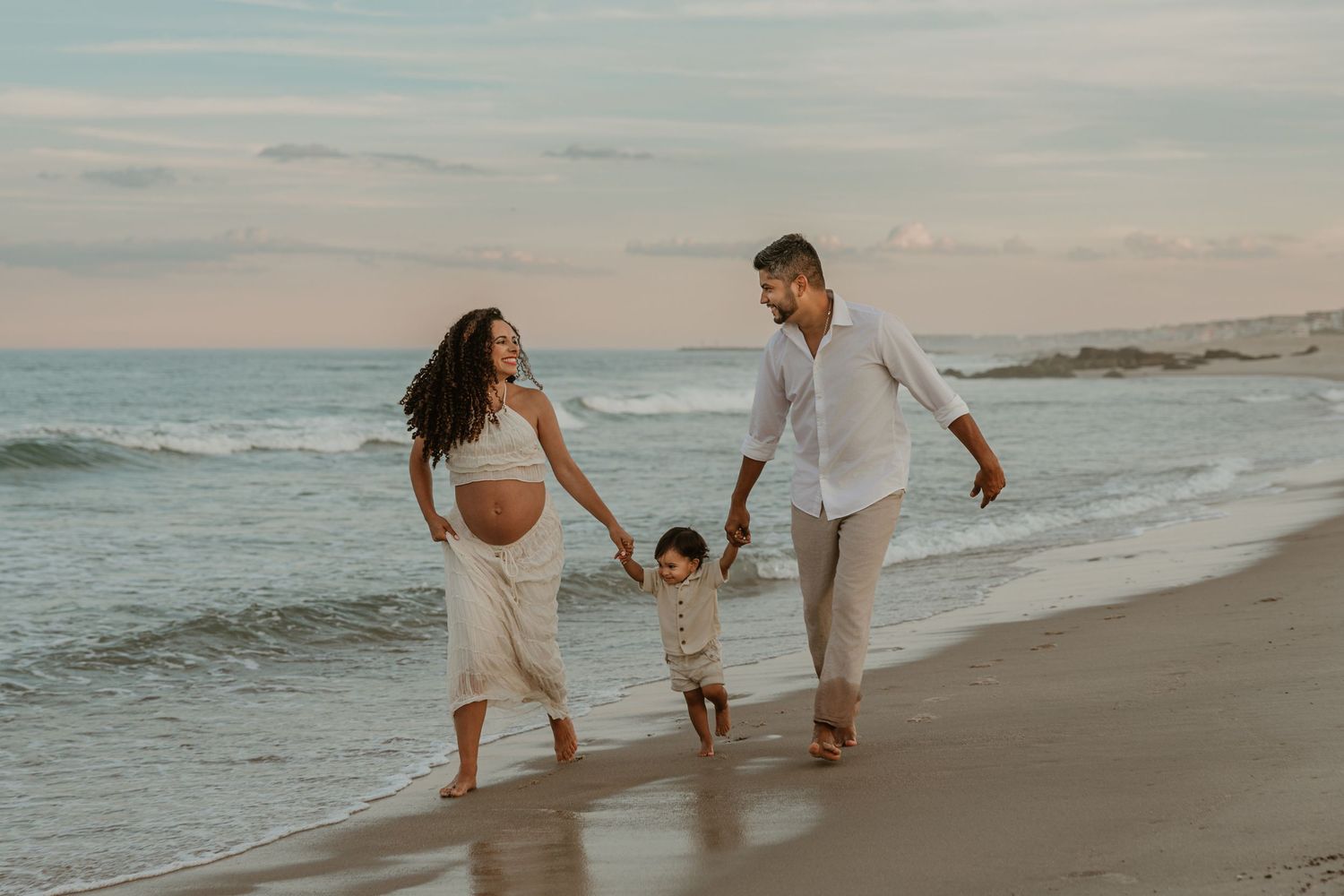 Pregnant woman and man walk on beach at sunset holding toddler's hands between them near ocean waves.