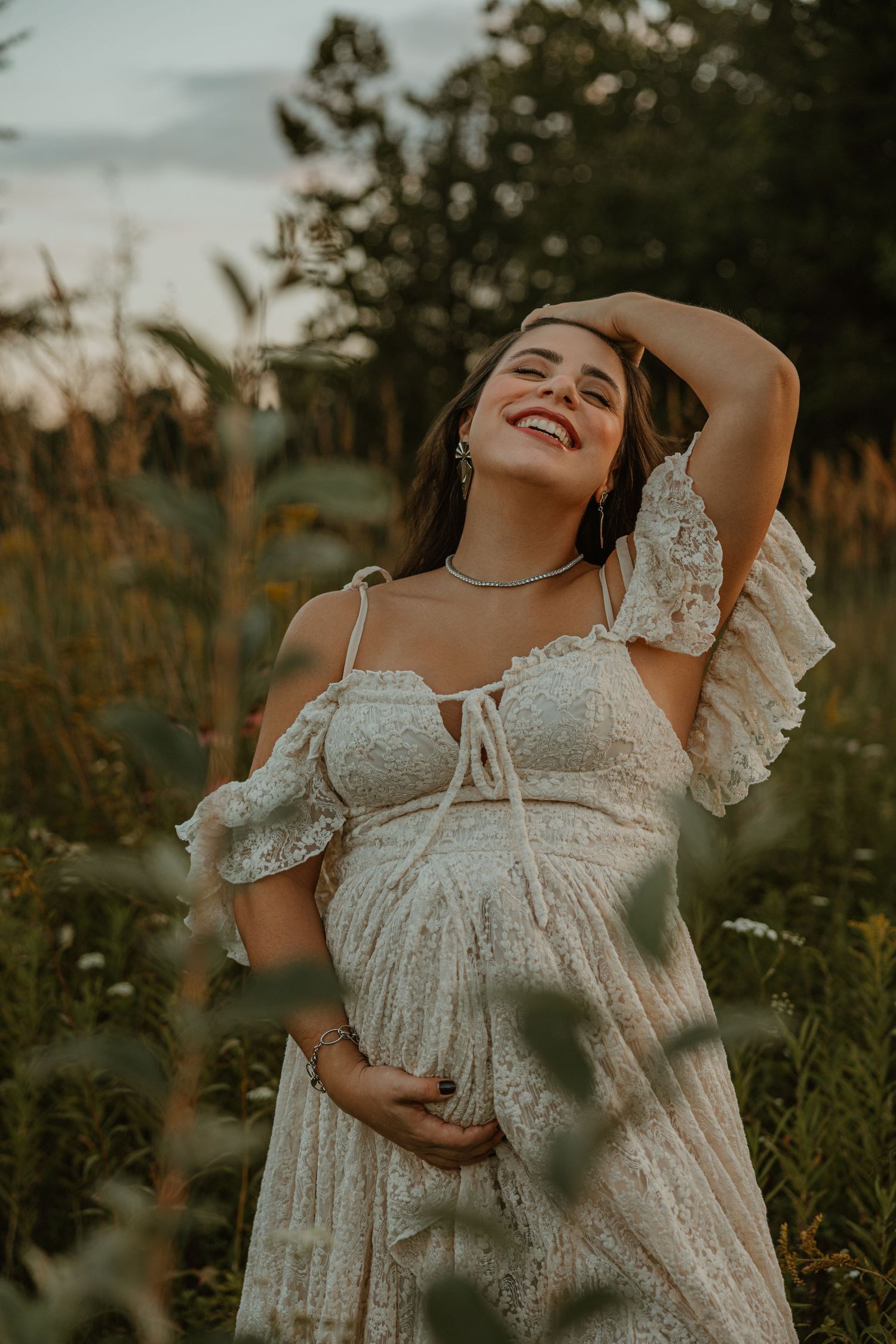 Smiling pregnant woman in white lace boho dress holding belly in golden hour field.