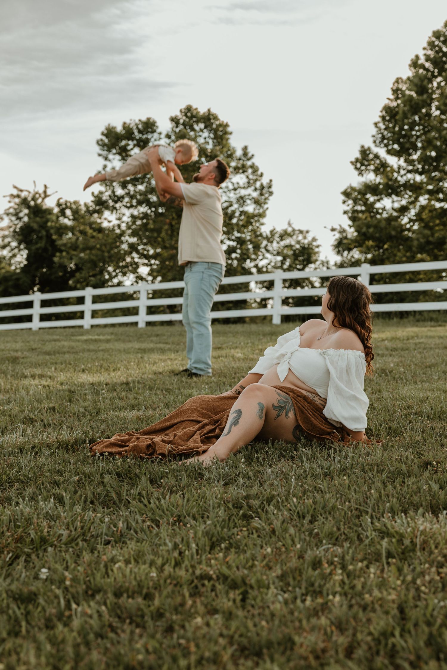 Pregnant woman sits on grass while man lifts baby in air during outdoor family maternity photo session.