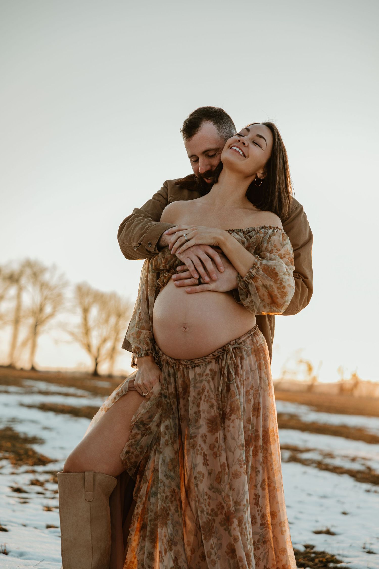 Intimate maternity photo of couple embracing in a floral dress against snowy landscape at sunset.