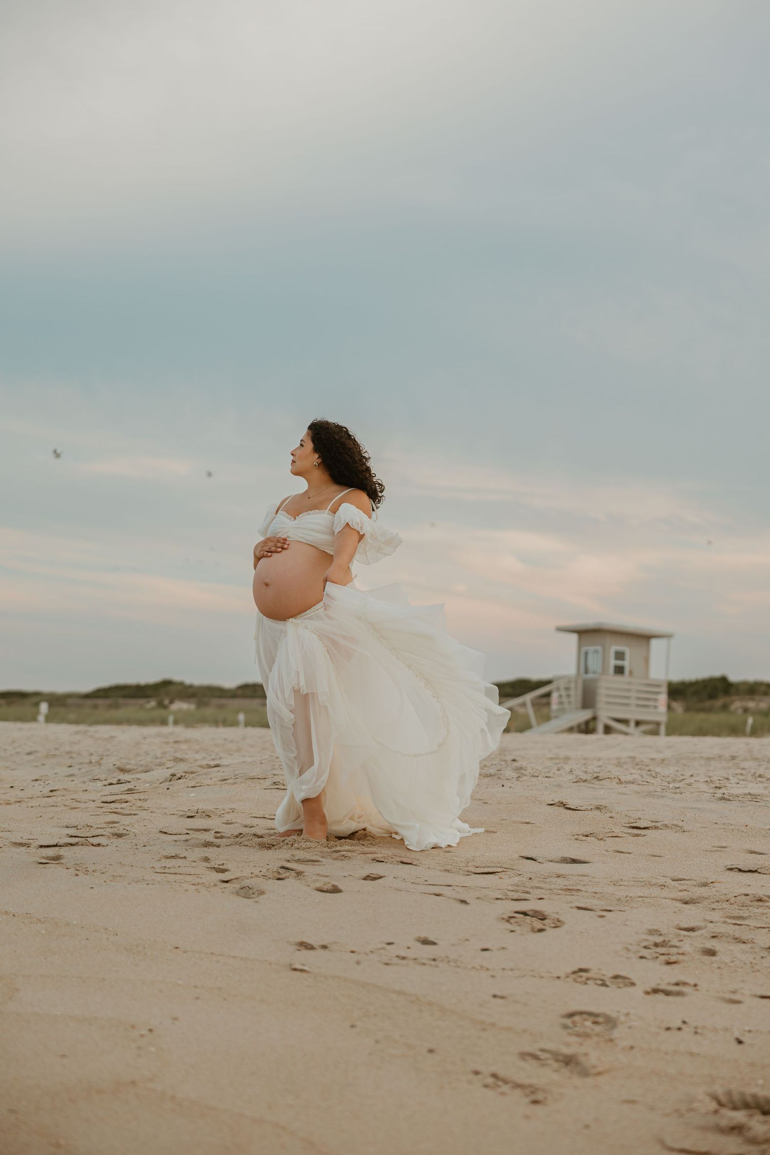 Pregnant woman in flowing white off-shoulder gown poses on sandy beach, cradling her belly, with a lifeguard tower in background.
