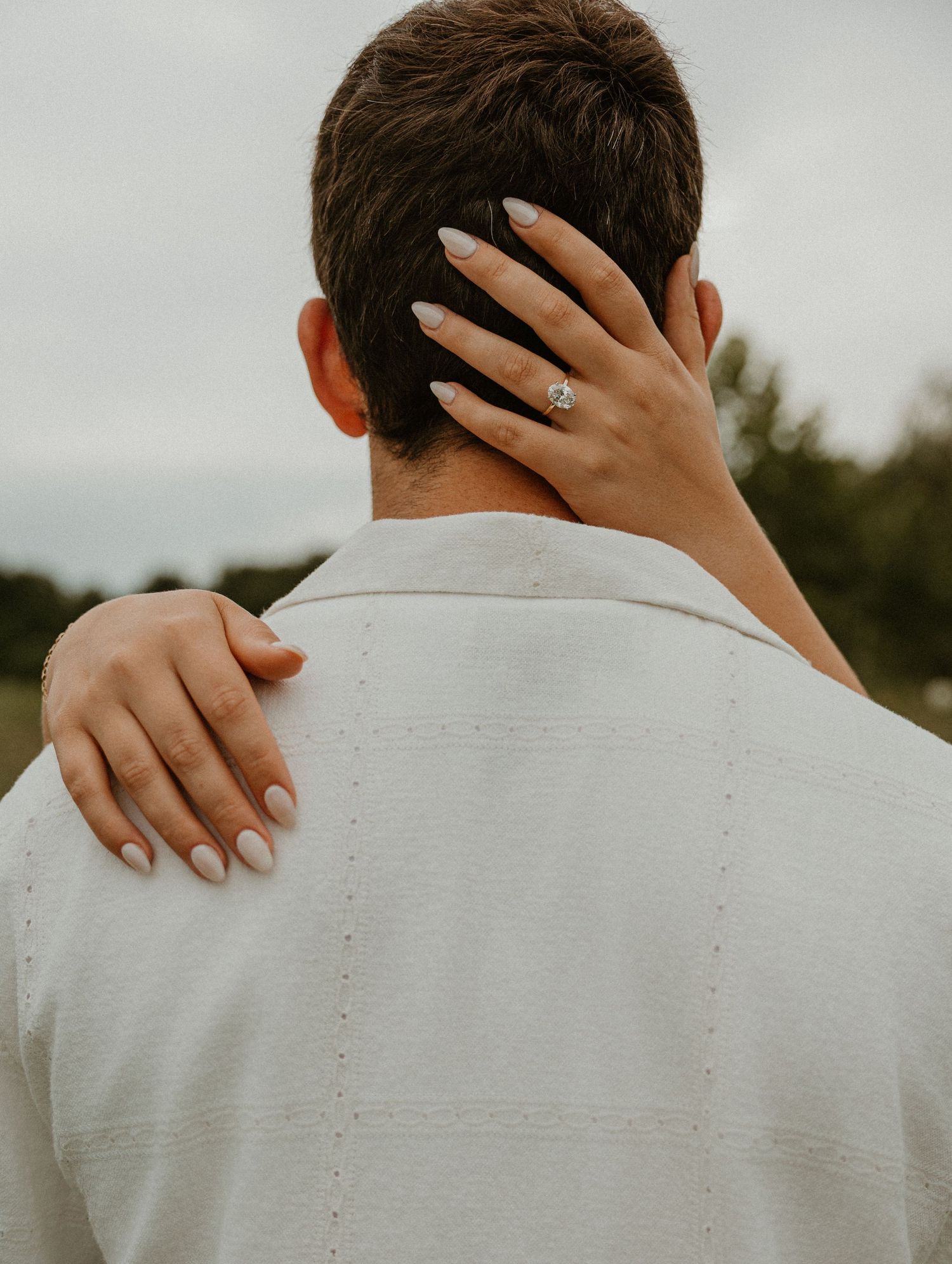 Woman with engagement ring rests hands on man's shoulders from behind, both facing away outdoors.
