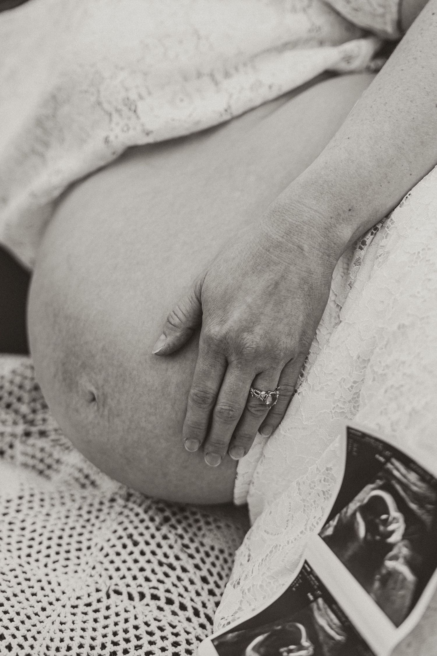Black and white close-up of pregnant woman's belly with hand resting on it and ultrasound photos nearby.