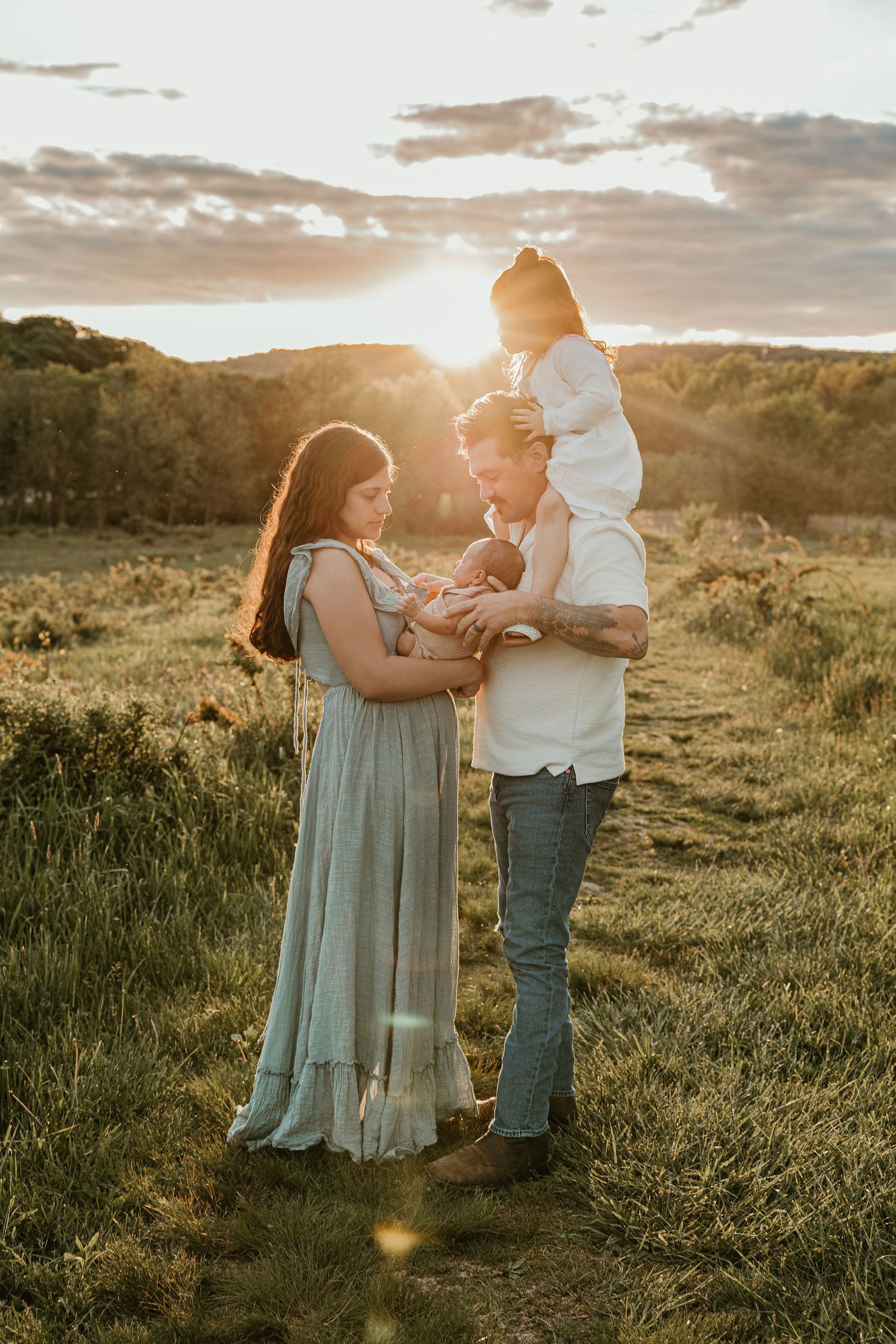 Family of four poses at golden hour in a field, parents holding newborn and toddler on shoulders.