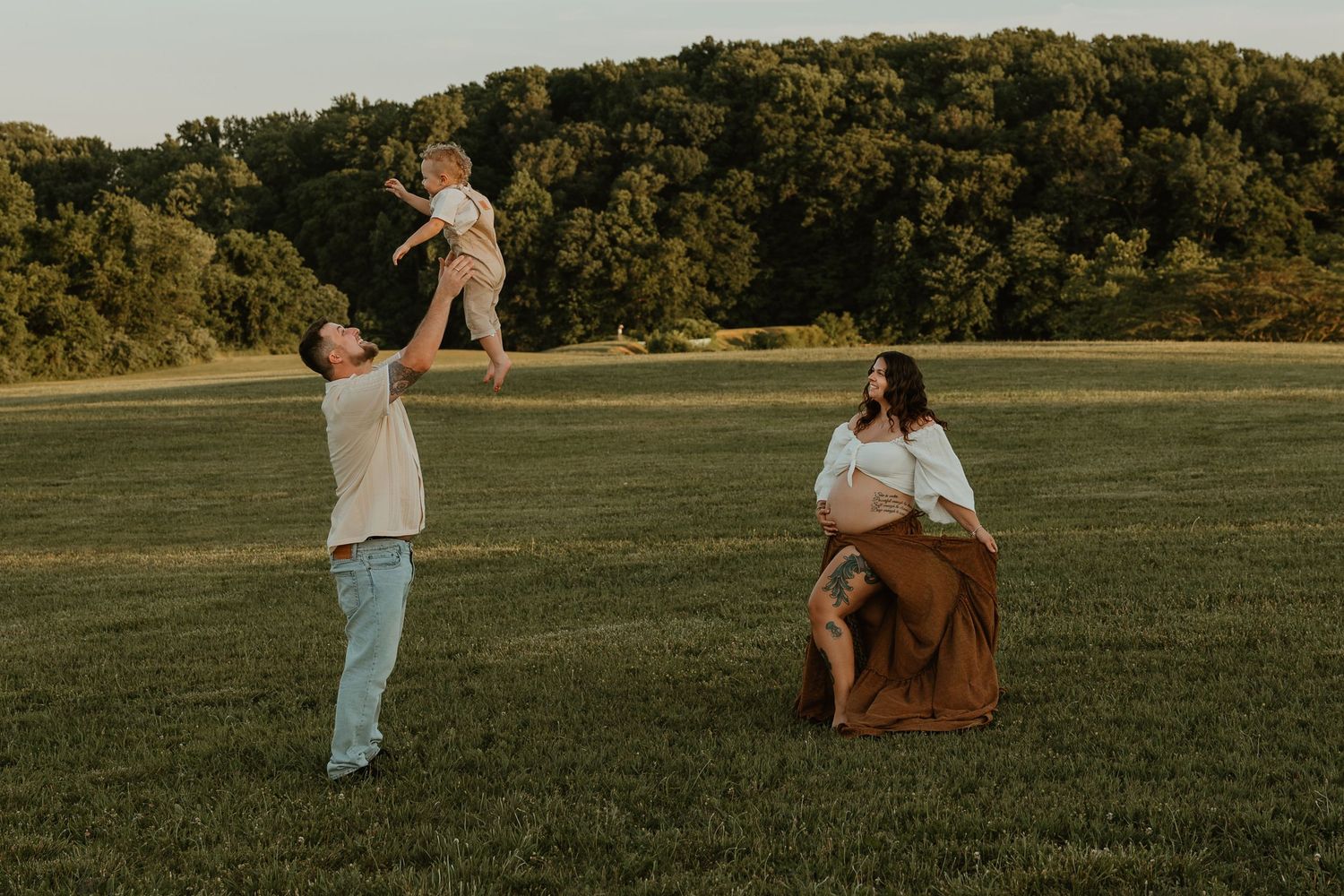 Pregnant woman watches husband toss toddler in air during golden hour family maternity photo session in green field.