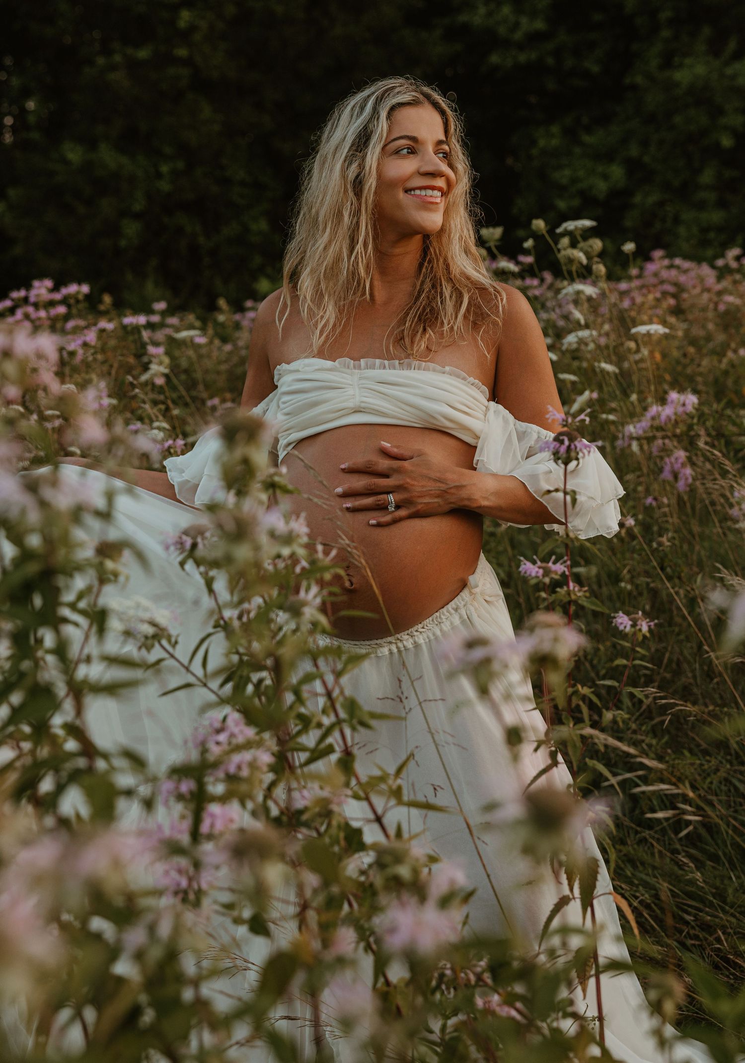 Smiling pregnant woman in white off-shoulder outfit cradling baby bump in a wildflower meadow.